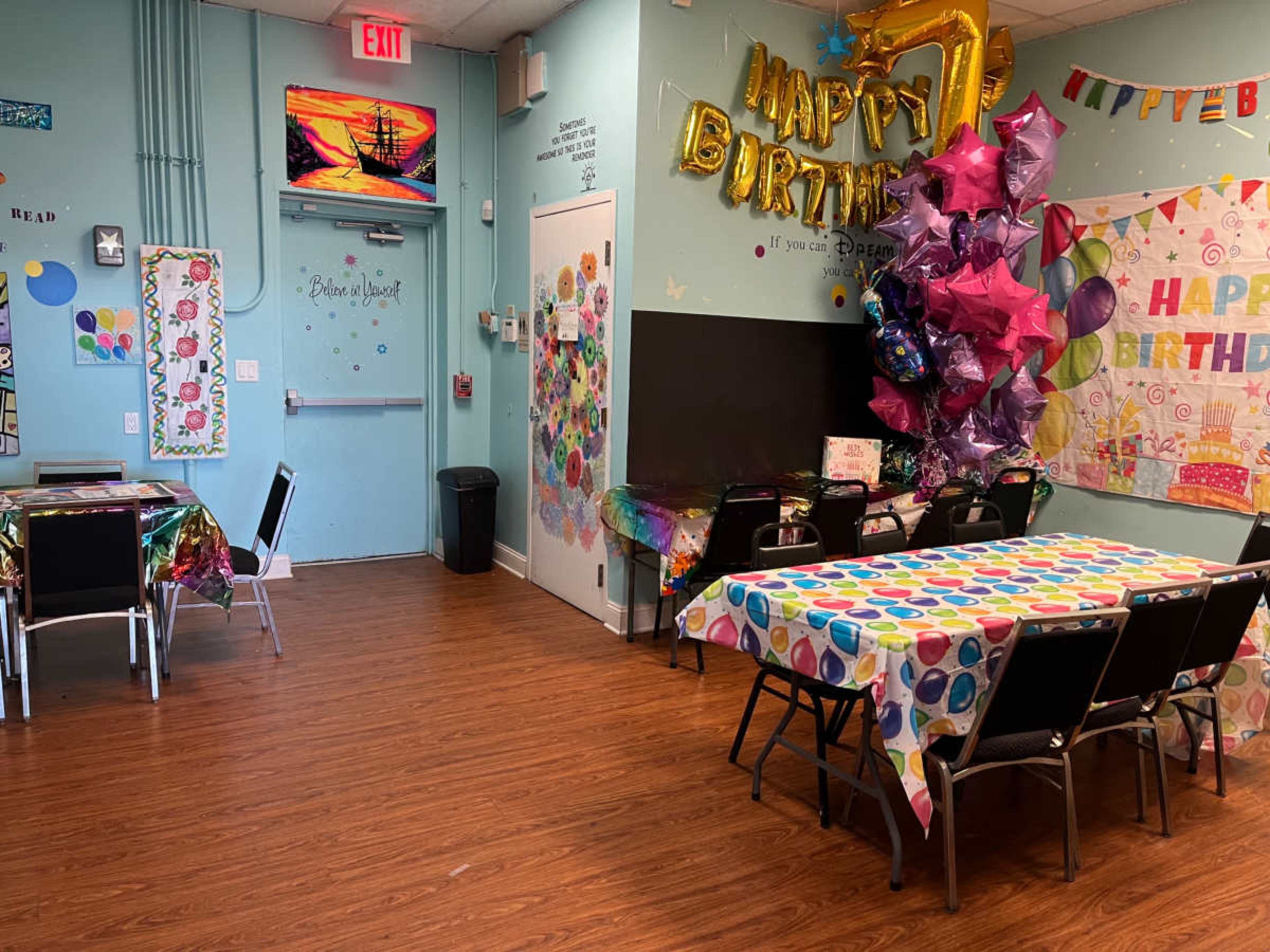 A brightly decorated room with colorful tables set for a birthday celebration, featuring balloons, streamers, and a "Happy Birthday" banner.