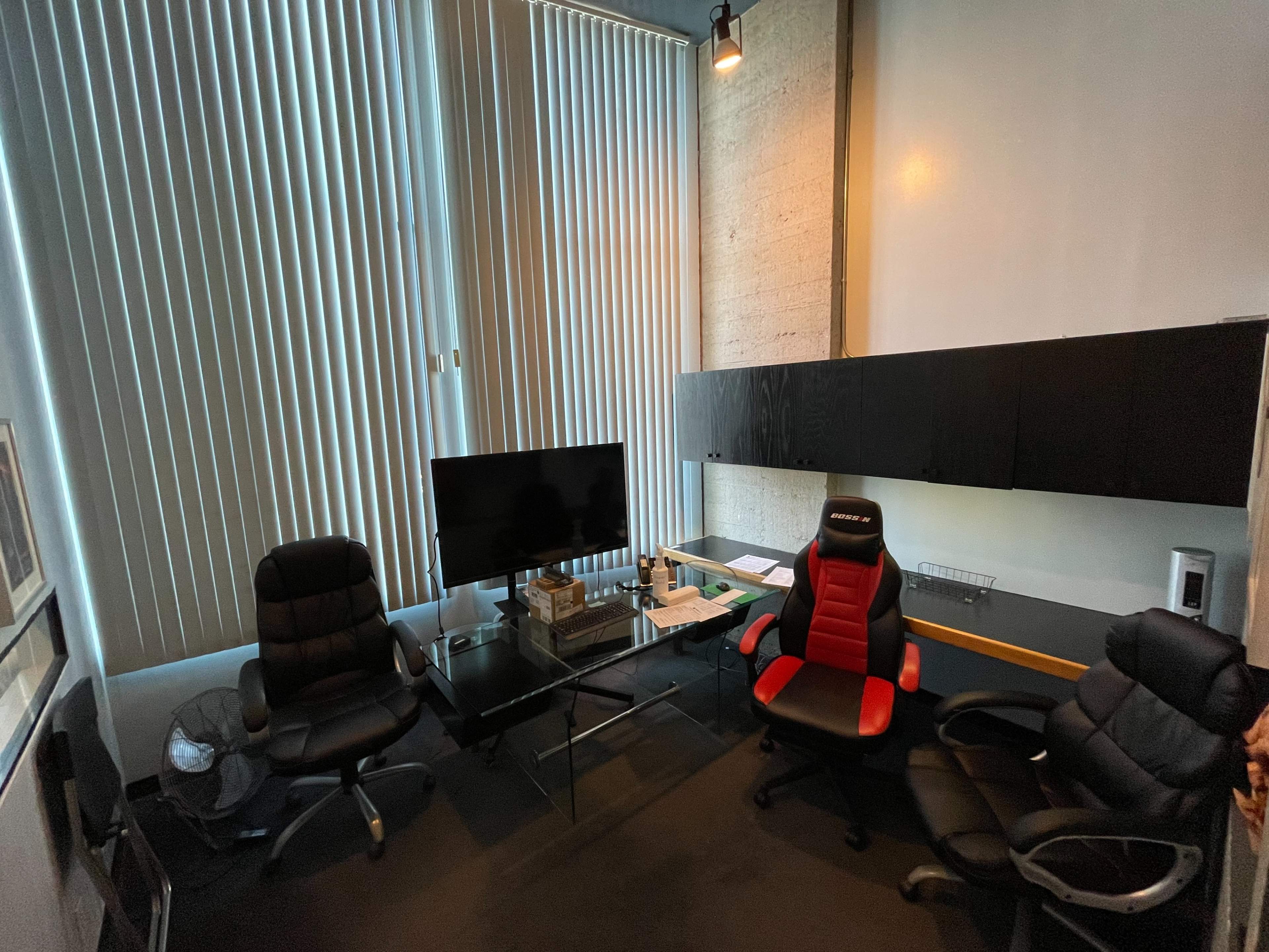 The image shows a small office space with two black office chairs and one red gaming chair, a glass table, a television, and large vertical blinds allowing natural light.