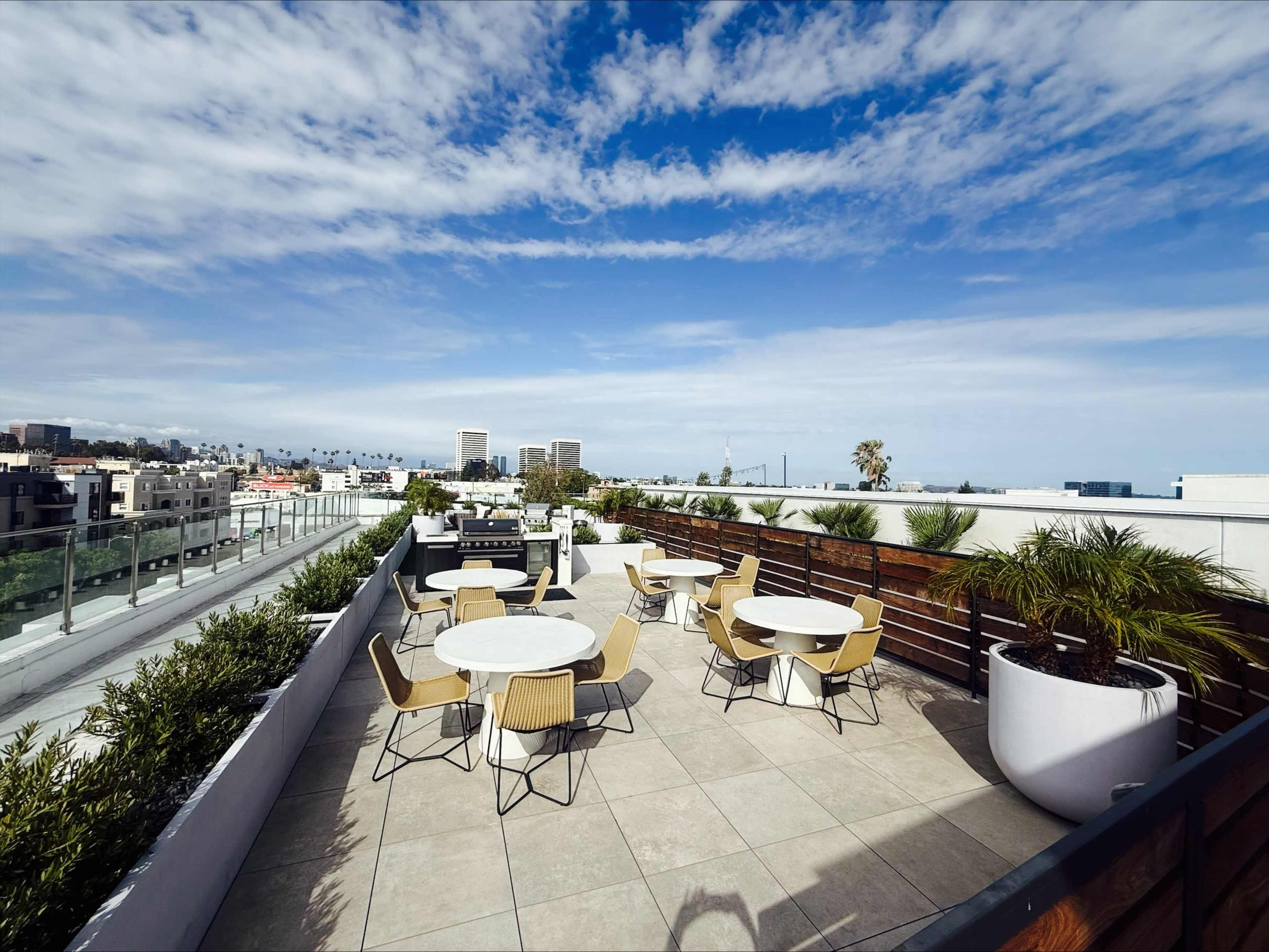 The image shows a rooftop terrace featuring several round tables and chairs, with a view of buildings and palm trees under a partly cloudy sky.
