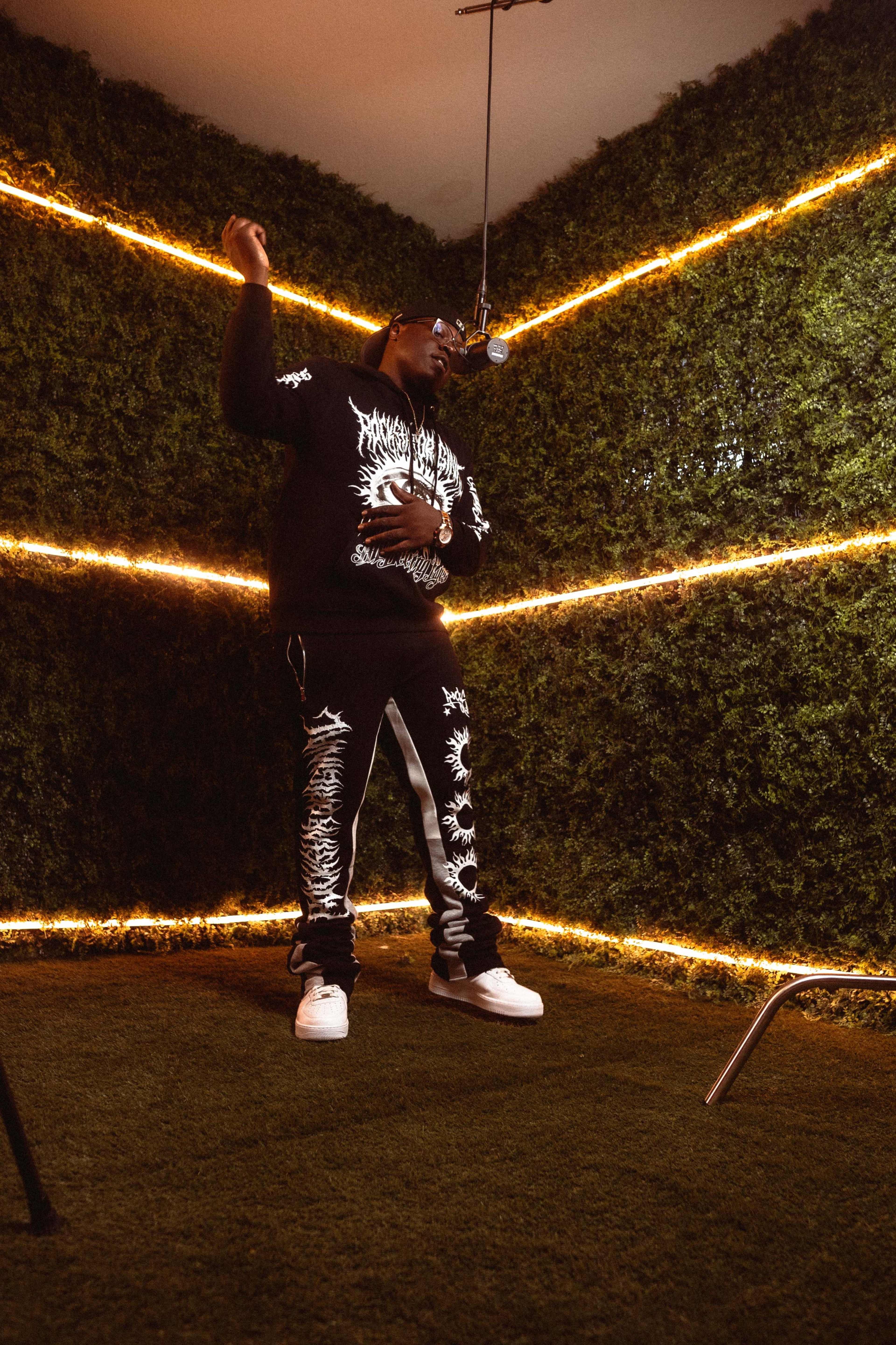 A person stands in a dimly lit room with a microphone, surrounded by green foliage and illuminated by string lights.