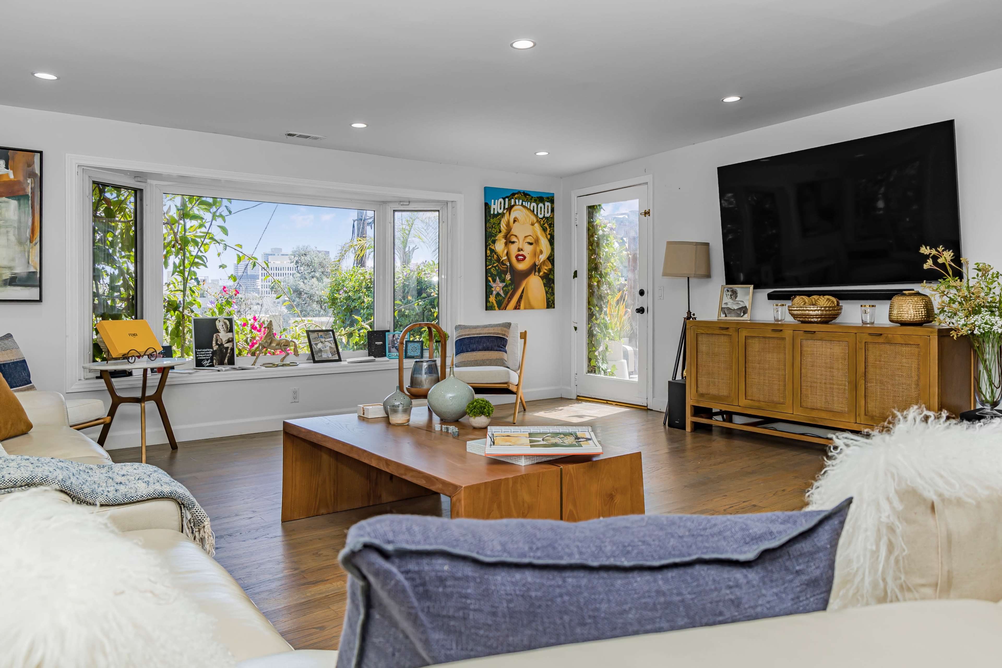 A bright living room features a large window with greenery outside, a wooden coffee table in the center, and a television mounted on the wall.