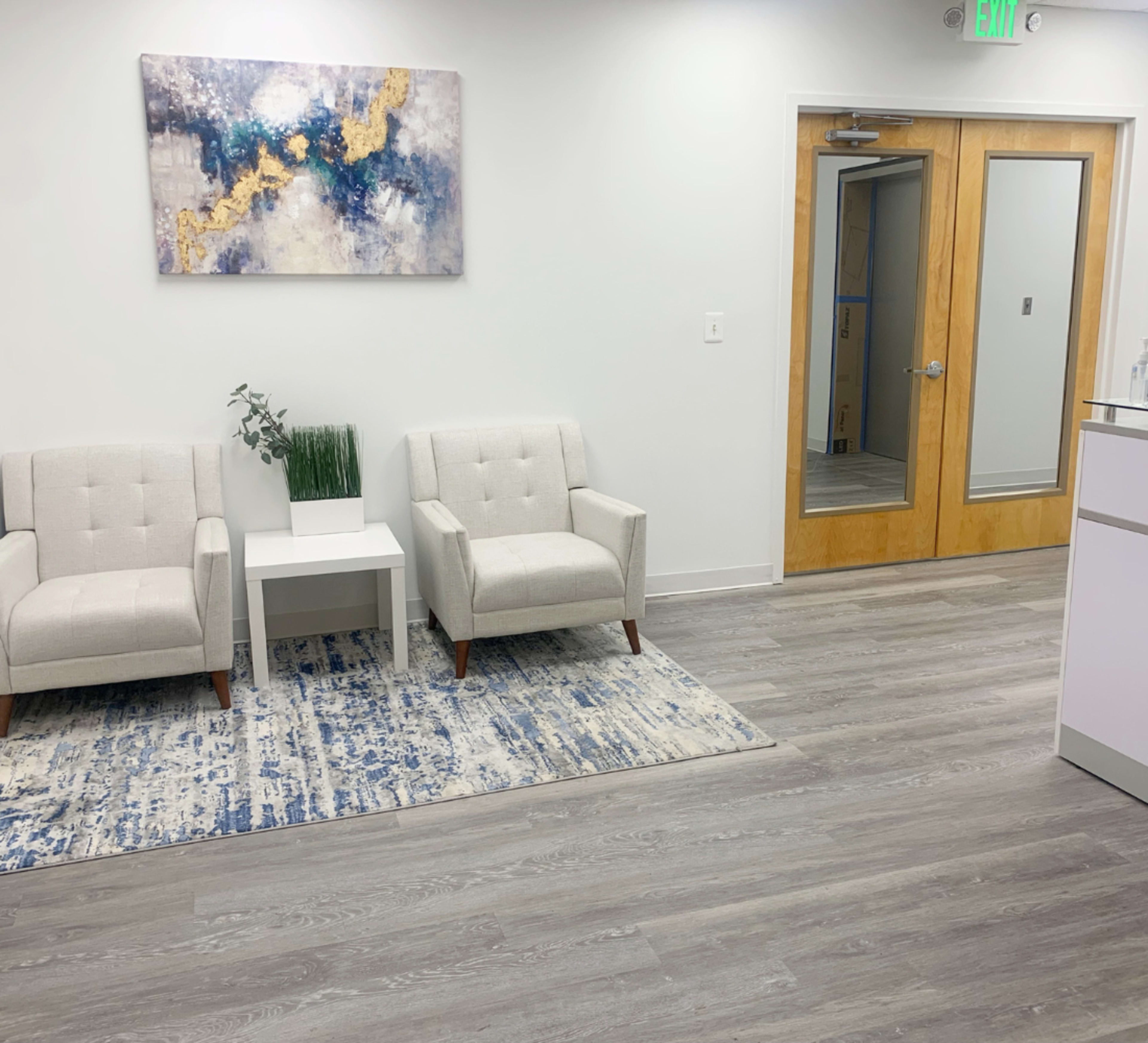 The image shows a waiting area with two light-colored armchairs, a small white side table, a green plant, and a modern art piece on the wall.
