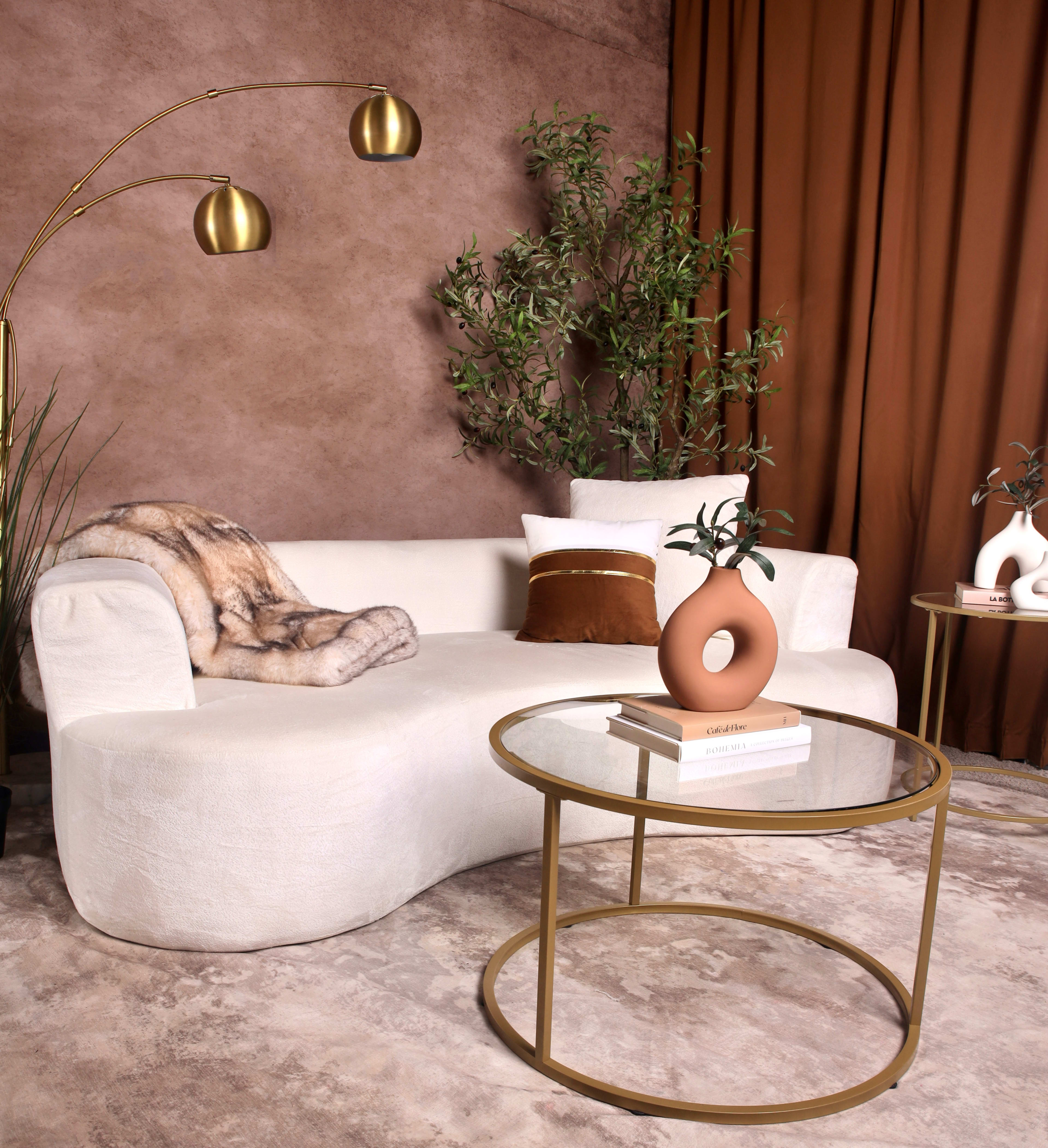 The image features a modern, curved white sofa with a fur throw, a circular glass coffee table, and decorative items set against a backdrop of brown curtains and a textured wall.