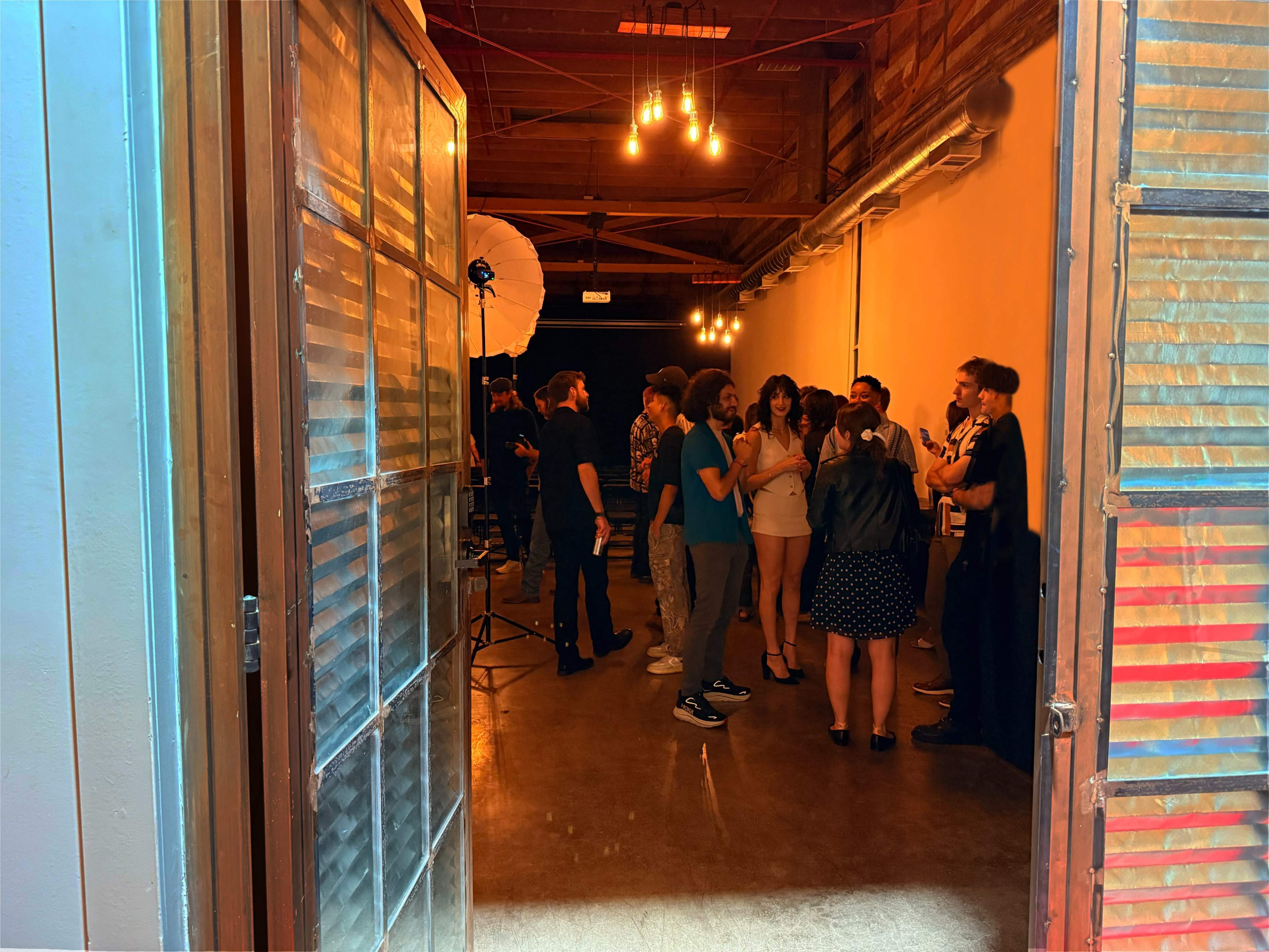 A group of people are gathered inside a well-lit room with exposed beams and studio equipment set up for a photo shoot.
