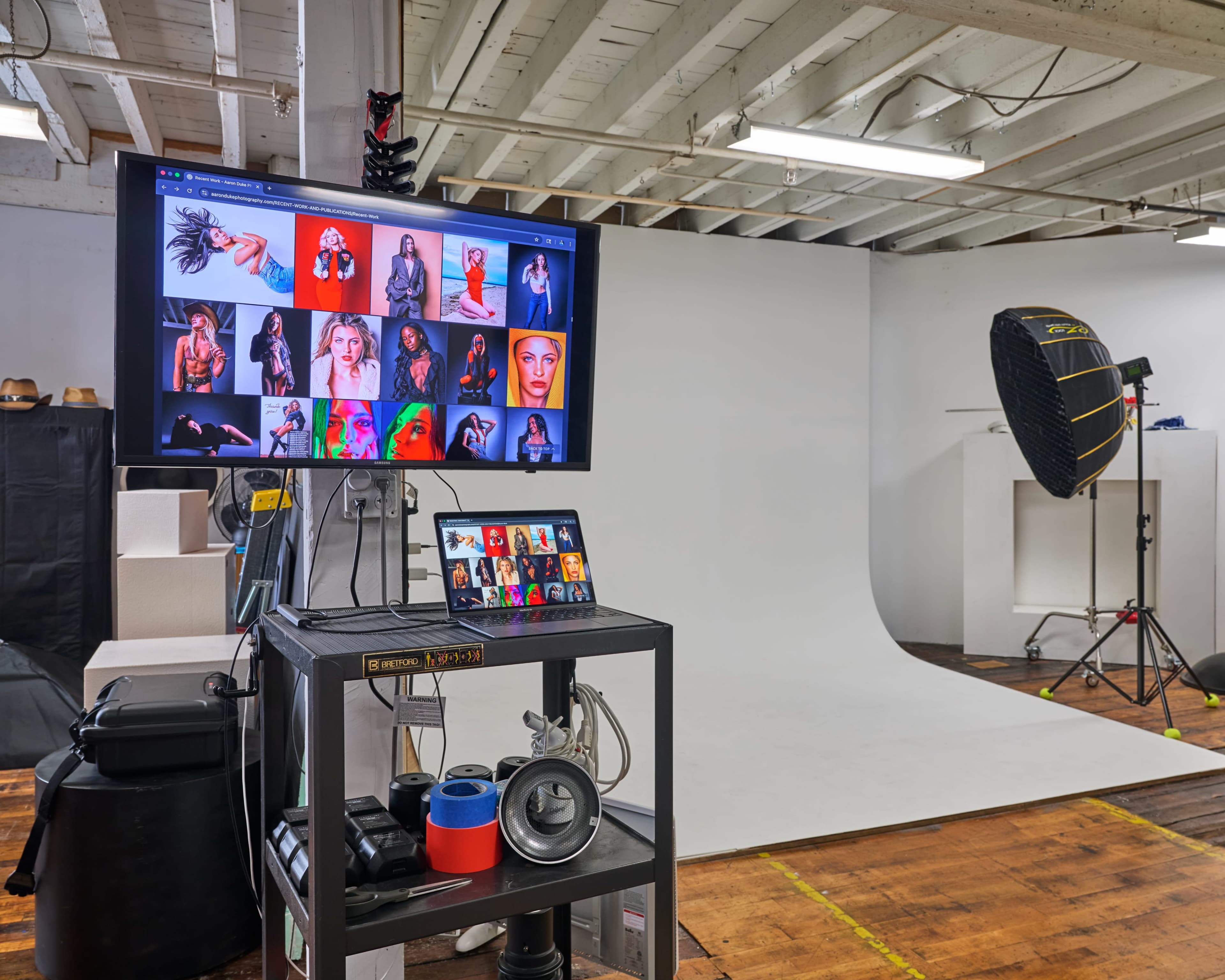 The image shows a photography studio setup with a monitor displaying various edited photos, a backdrop, lighting equipment, and a cart with photographic tools.