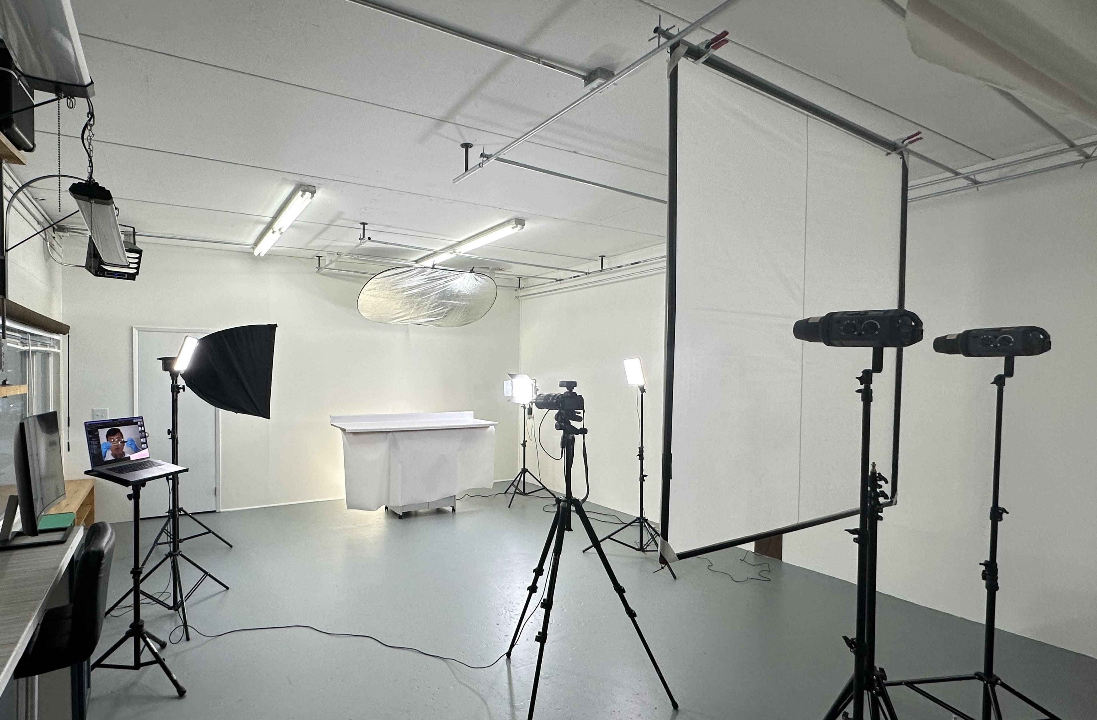 A photo studio setup features a white backdrop, multiple softbox lights, two cameras on tripods, and a table covered with a cloth.