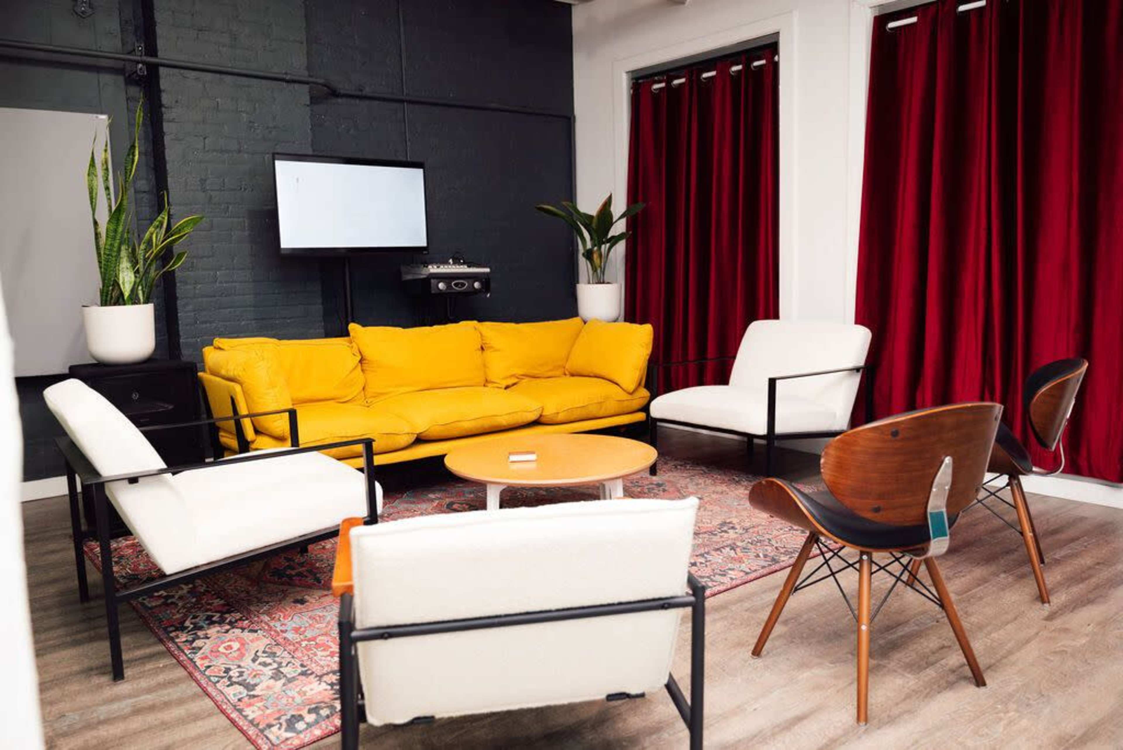 A modern seating area with a bright yellow couch, white chairs, a round table, and red curtains in a room featuring plants and a television on the wall.