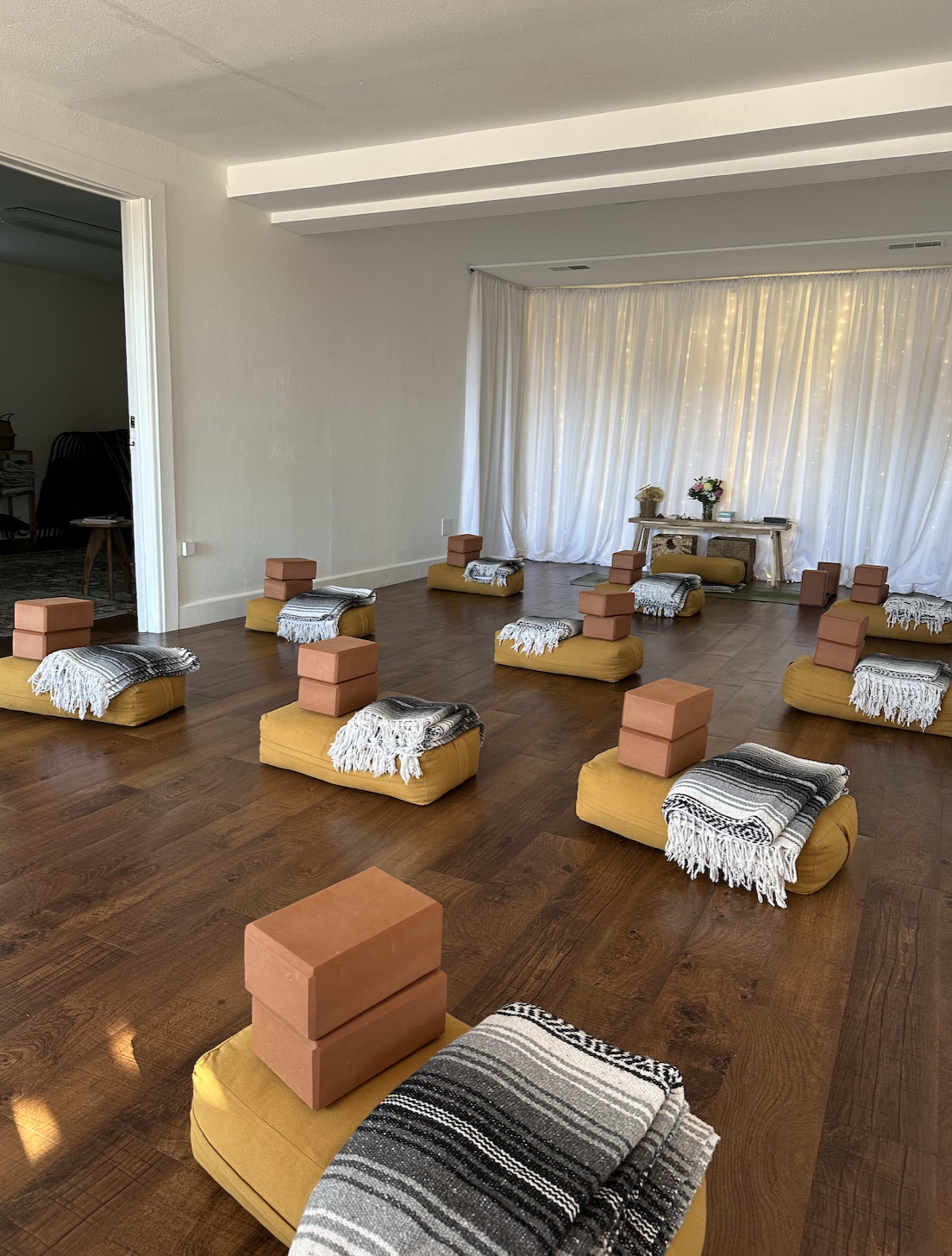 The image shows a room arranged for a yoga or meditation session, featuring mustard-colored seating cushions, yoga blocks, and blankets neatly organized on a wooden floor.