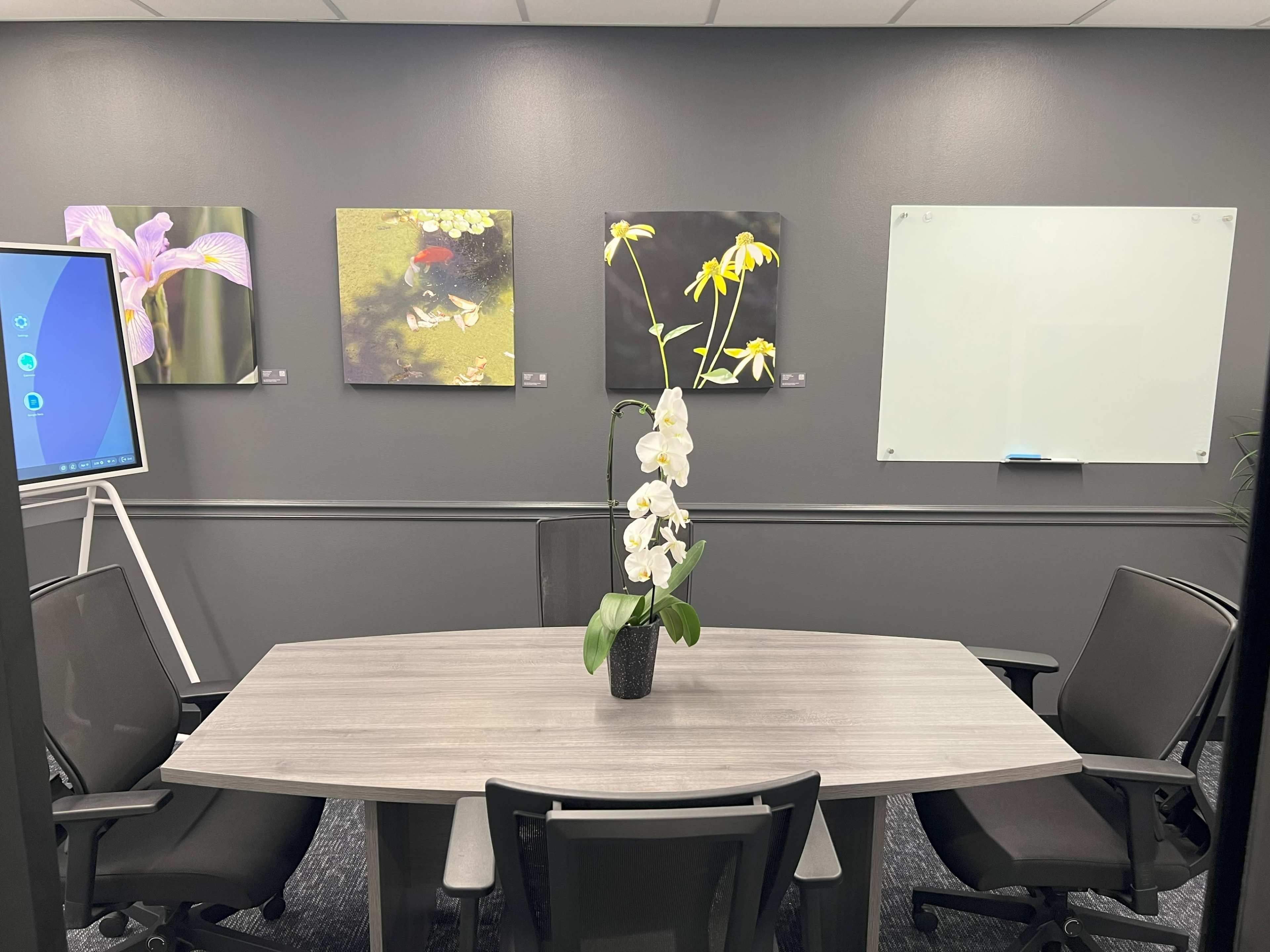 The image shows a conference room featuring a round table surrounded by chairs, with floral artwork on the gray walls and a whiteboard on one side.