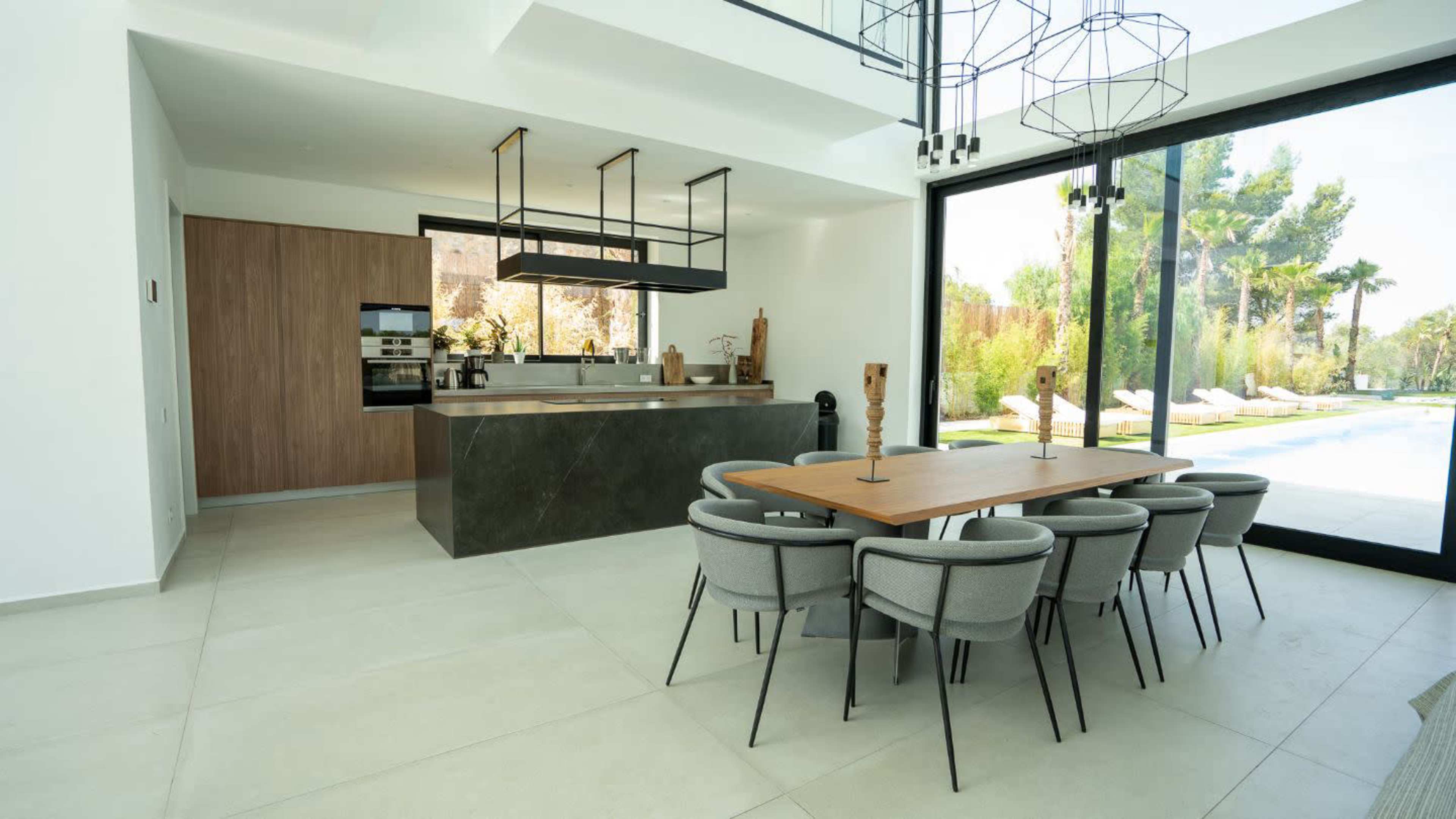 A modern kitchen and dining area featuring a large wooden table surrounded by gray upholstered chairs, with a sleek kitchen island and large windows overlooking an outdoor space.