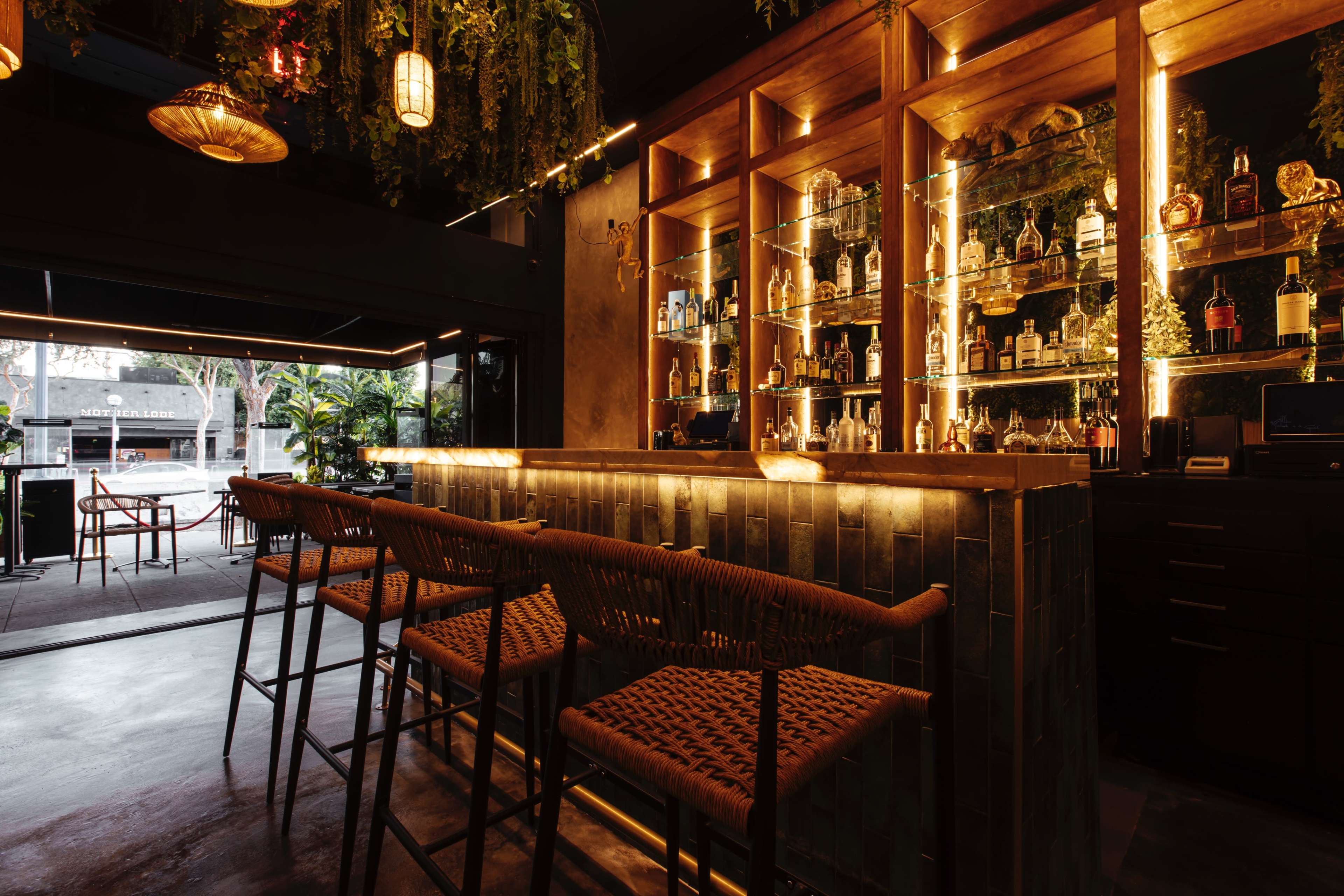 A stylish bar with wooden shelving displaying various bottles, illuminated by warm lighting, and surrounded by modern seating.