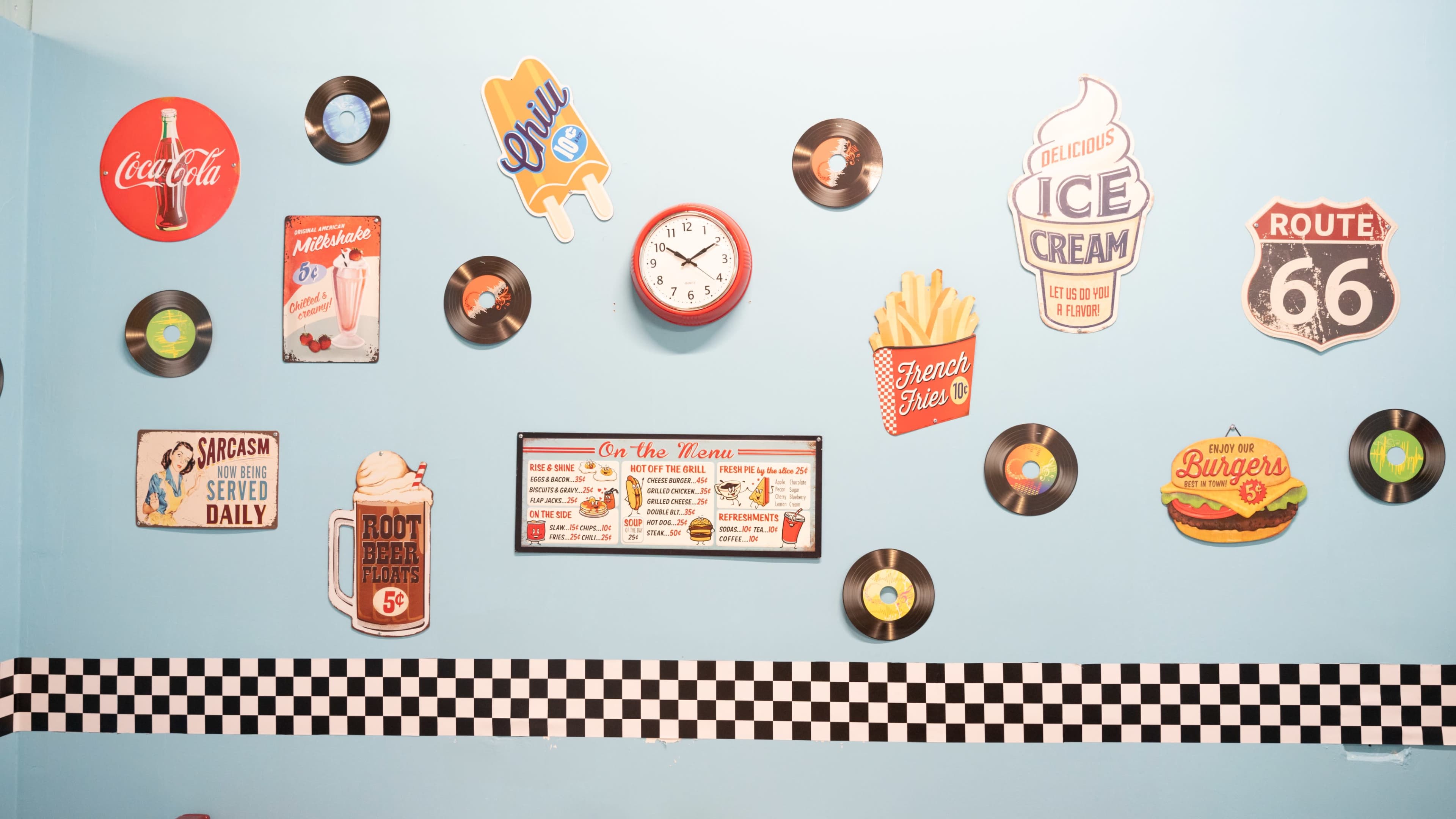 The image features a blue wall decorated with vintage signs and records, including advertisements for Coca-Cola, ice cream, and Route 66, alongside a clock and a menu board.