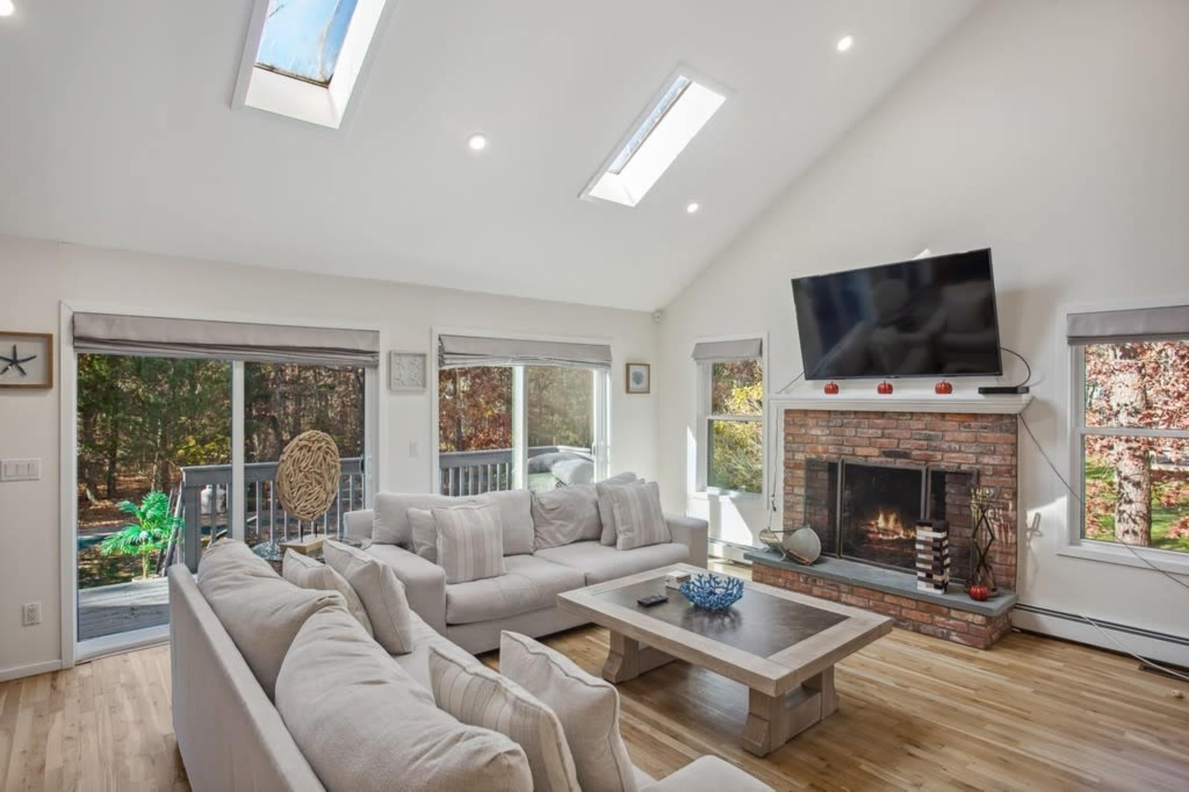 The image shows a modern living room with a large sectional sofa, a coffee table, and a flat-screen TV mounted above a brick fireplace, with skylights and windows letting in natural light.