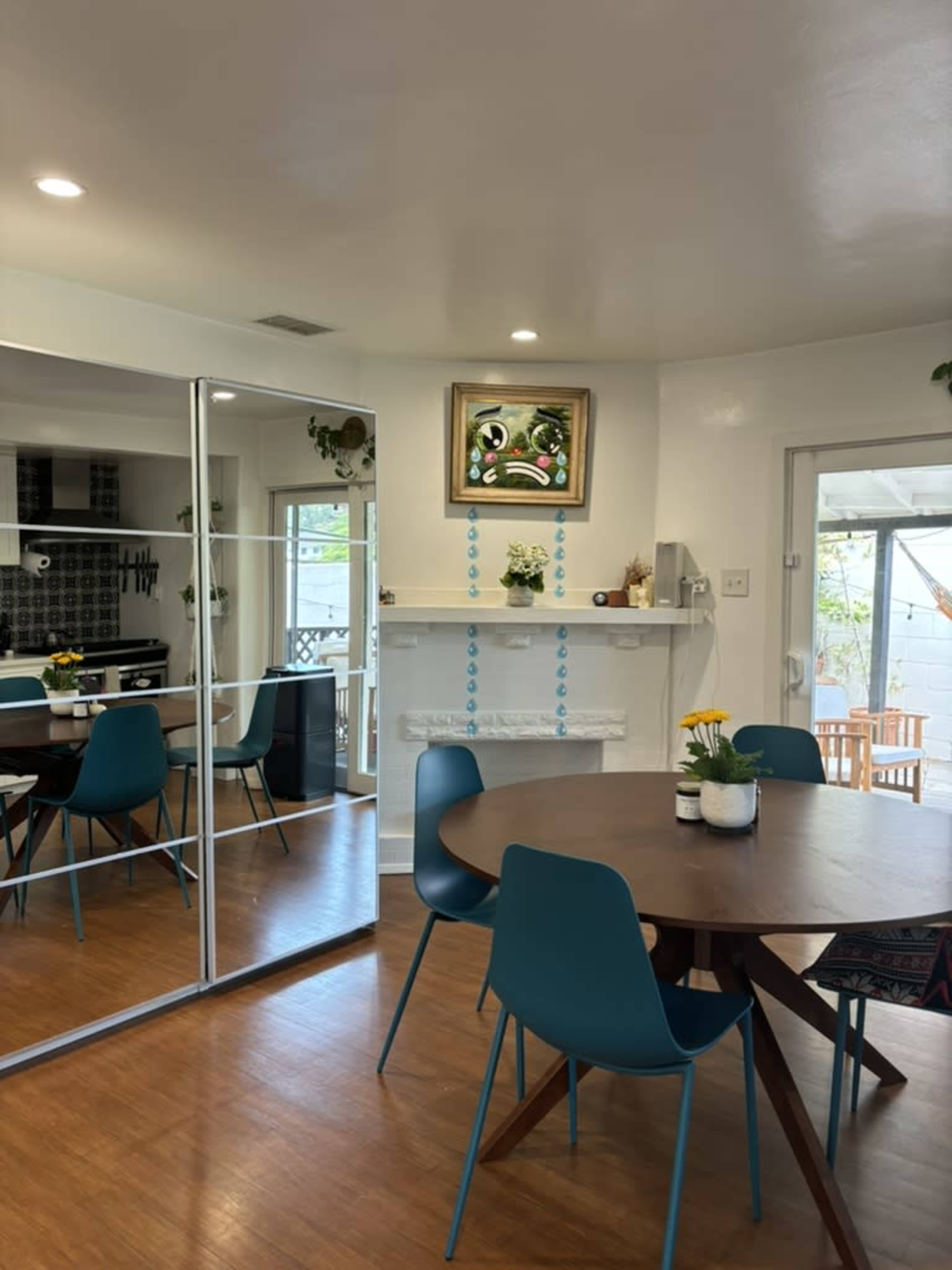 A dining area features a round wooden table with teal chairs, mirrored cabinets, and a colorful painting above a white fireplace.