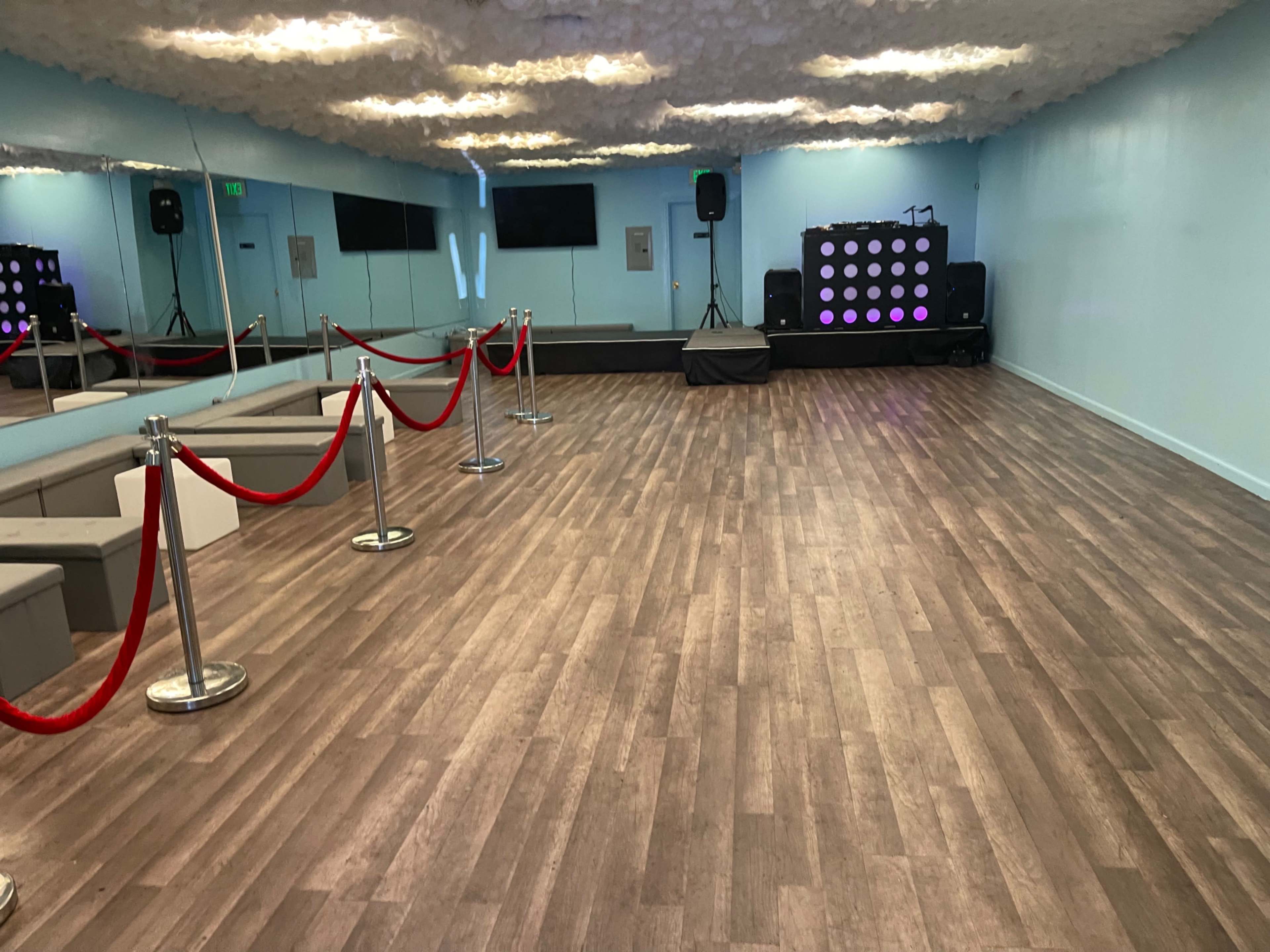 A spacious, empty event venue with a wooden floor, red ropes marking off an area, a stage with speakers, and a DJ setup in the background.