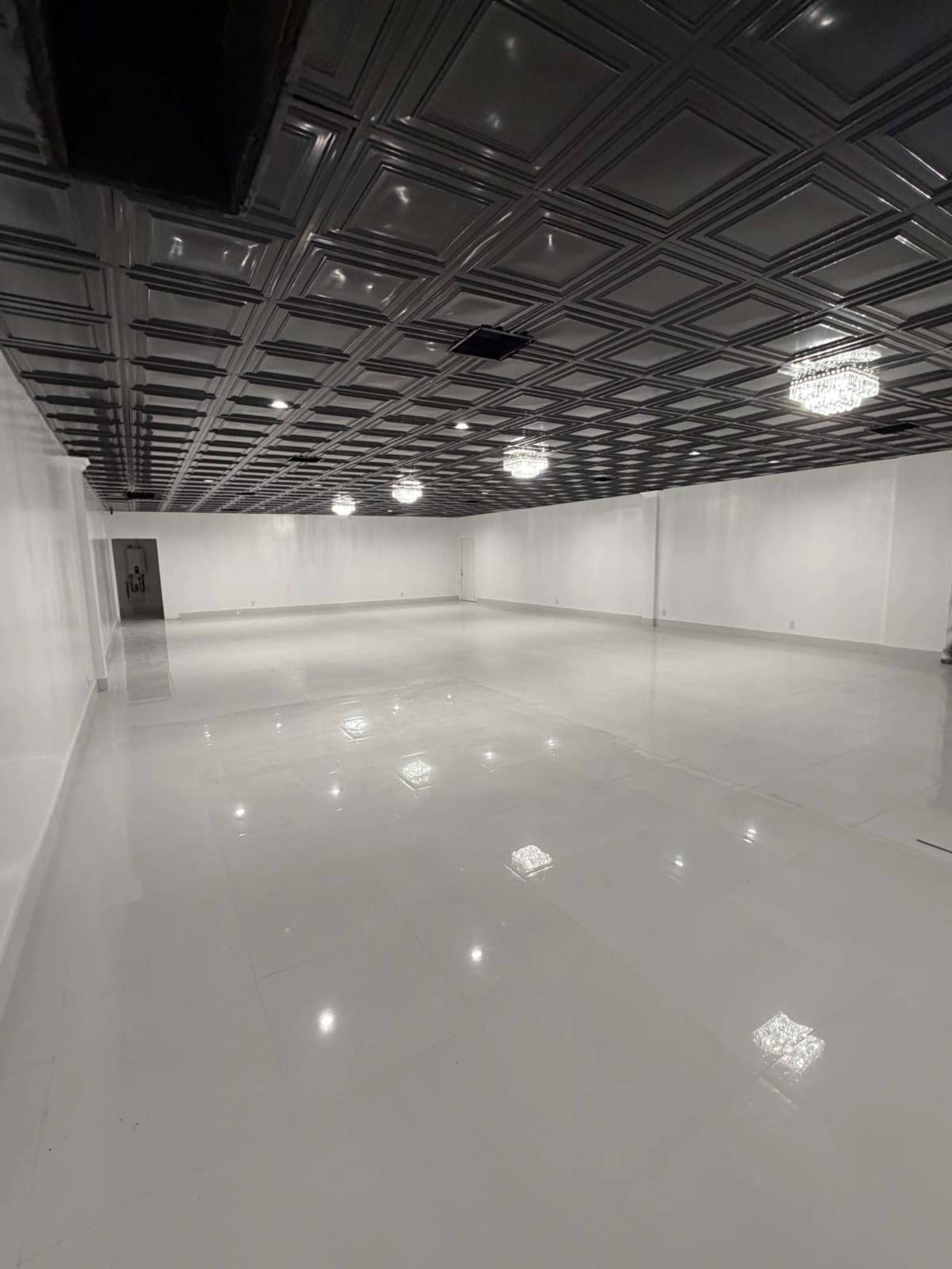 The image shows a large, empty room with glossy white tile flooring, a trey ceiling, and several hanging lights.