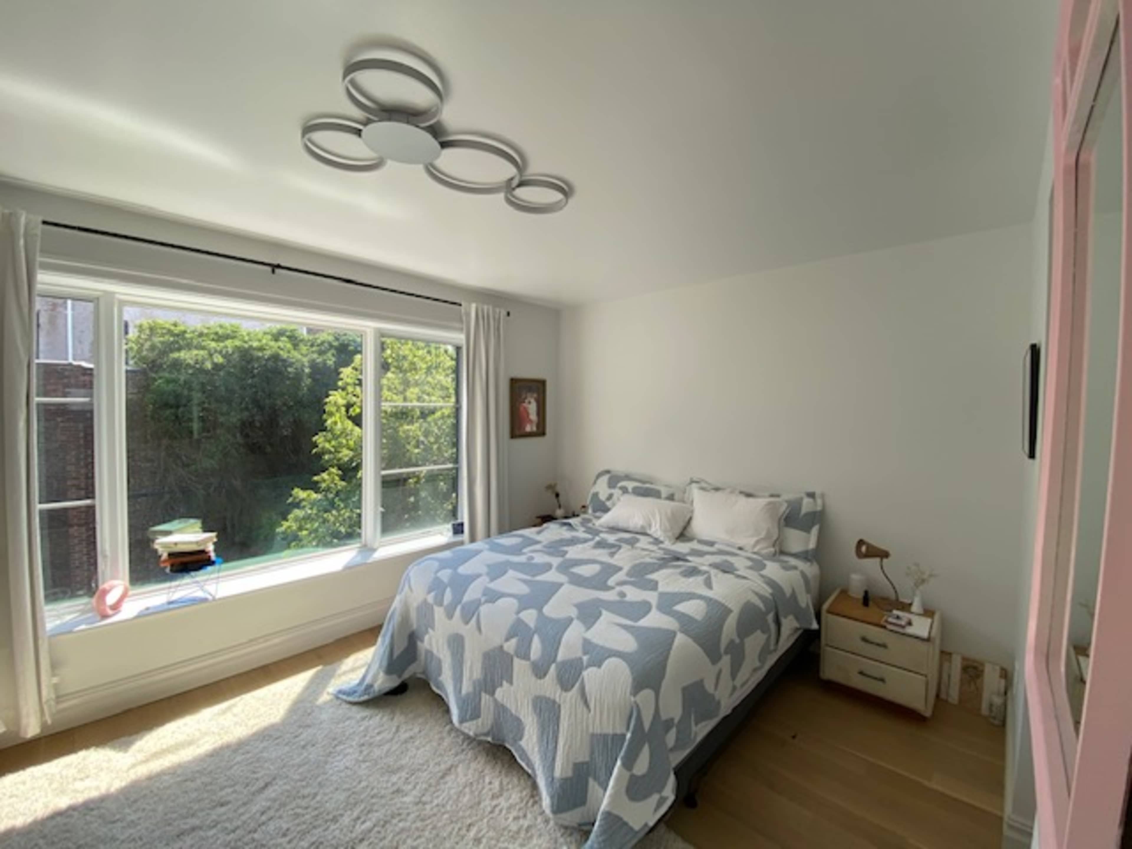 The image shows a bright bedroom featuring a bed with a blue and white quilt, a large window with green foliage outside, and a bedside table with a lamp and decorative items.