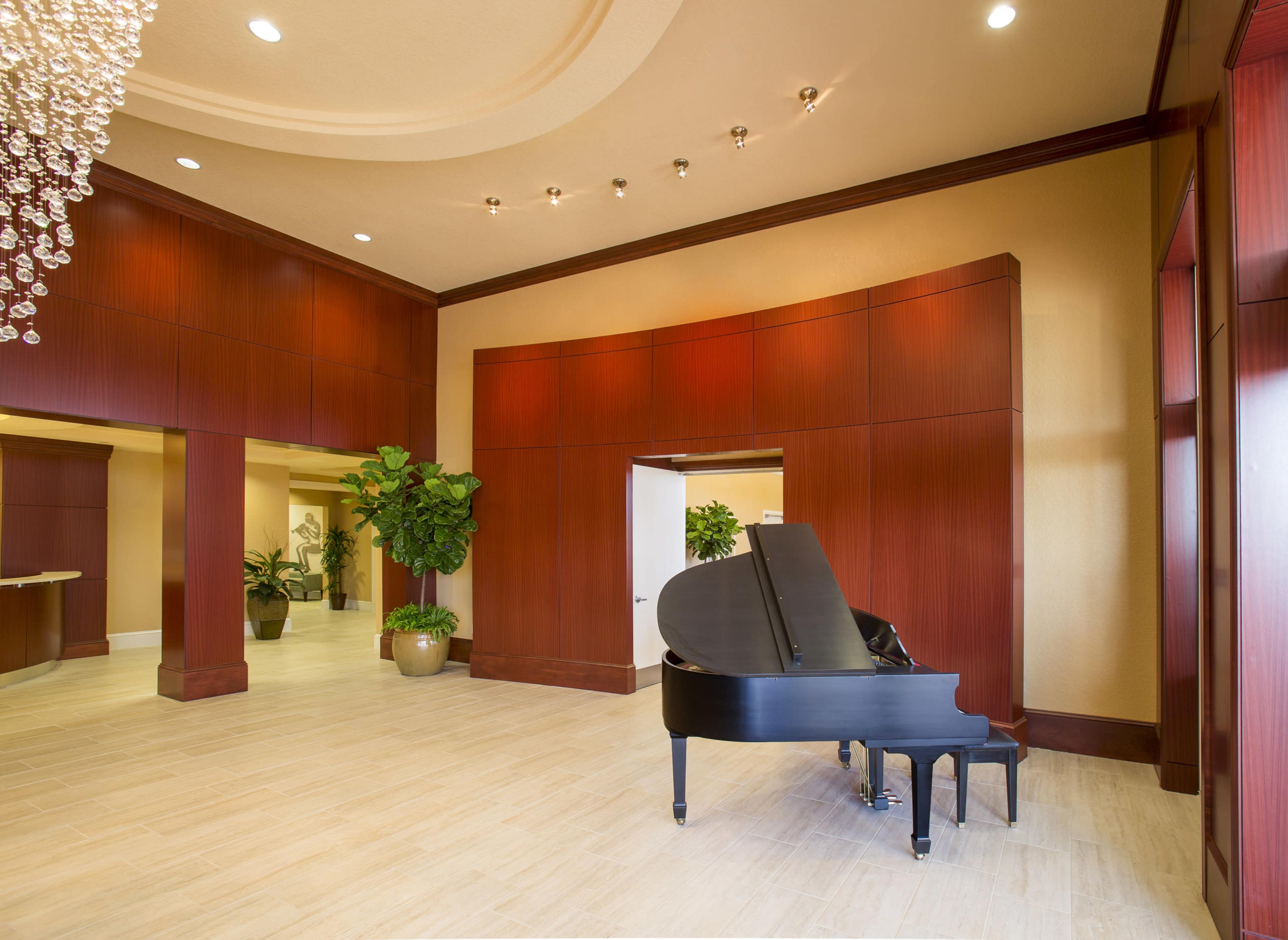 The image shows a spacious lobby featuring a black grand piano near a decorative chandelier and potted plants.
