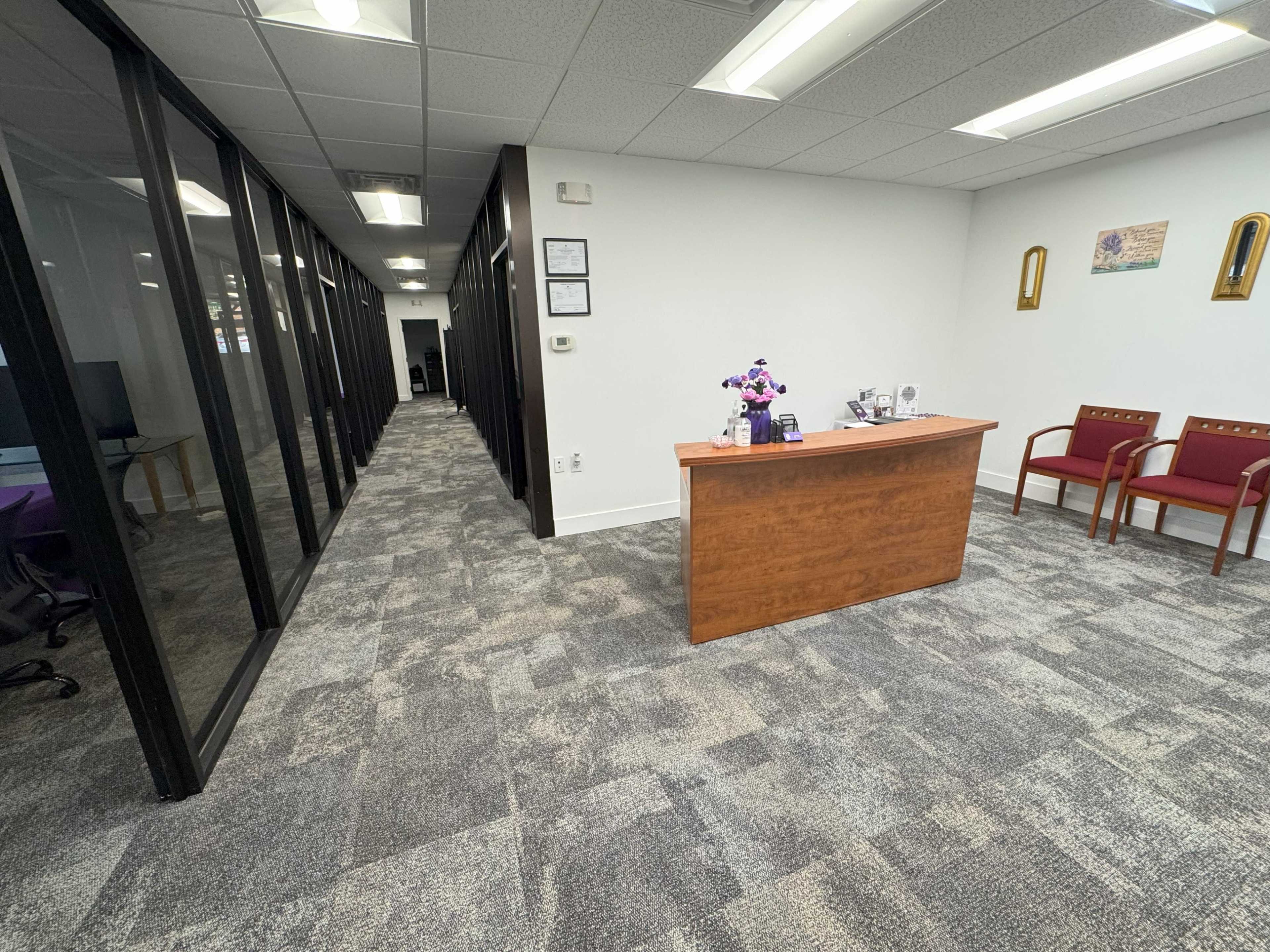 The image shows a modern office reception area with a wooden front desk, two waiting chairs, and a hallway leading to other office spaces.