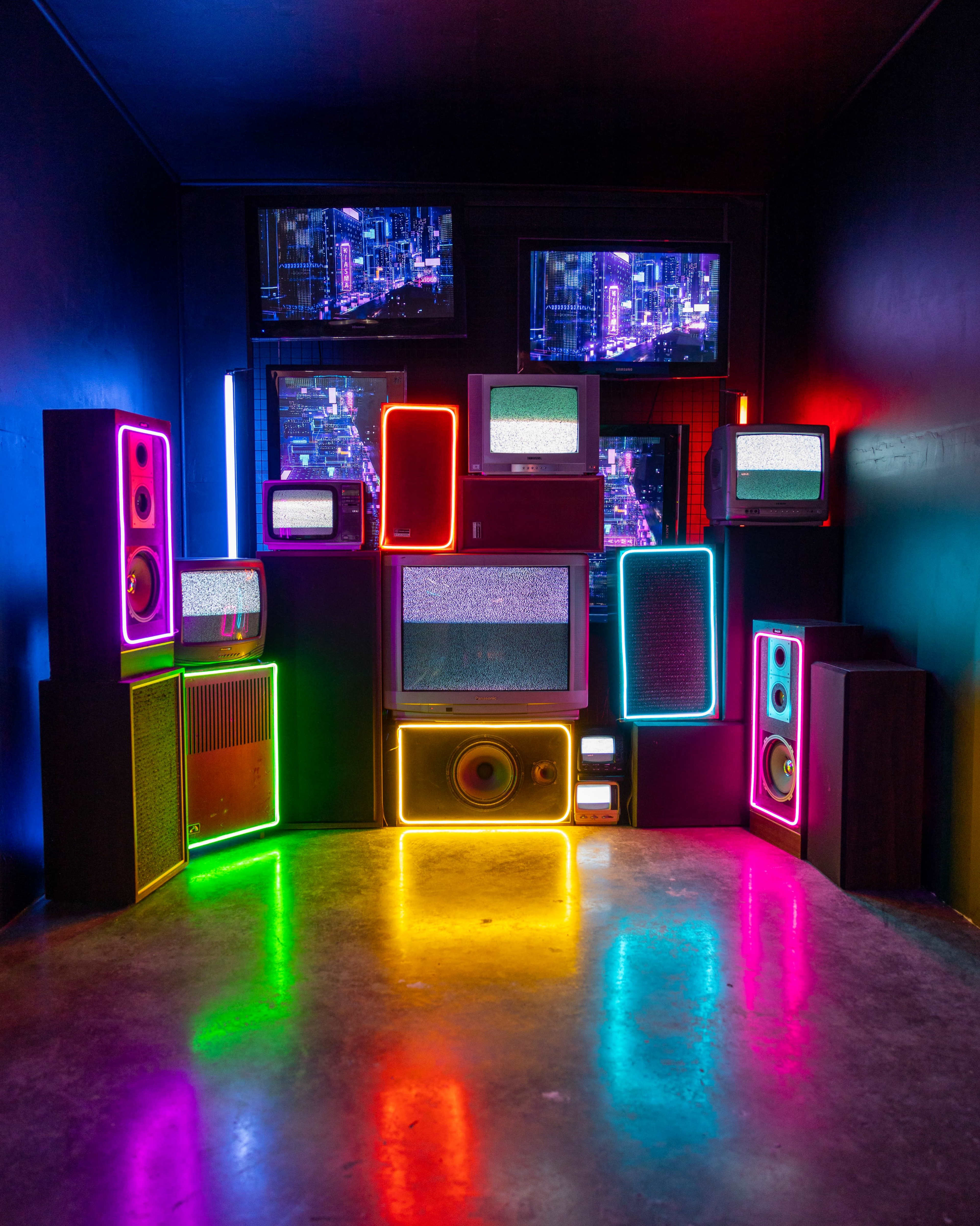 The scene features a collection of retro televisions and speakers illuminated by vibrant neon lights against a dark wall.