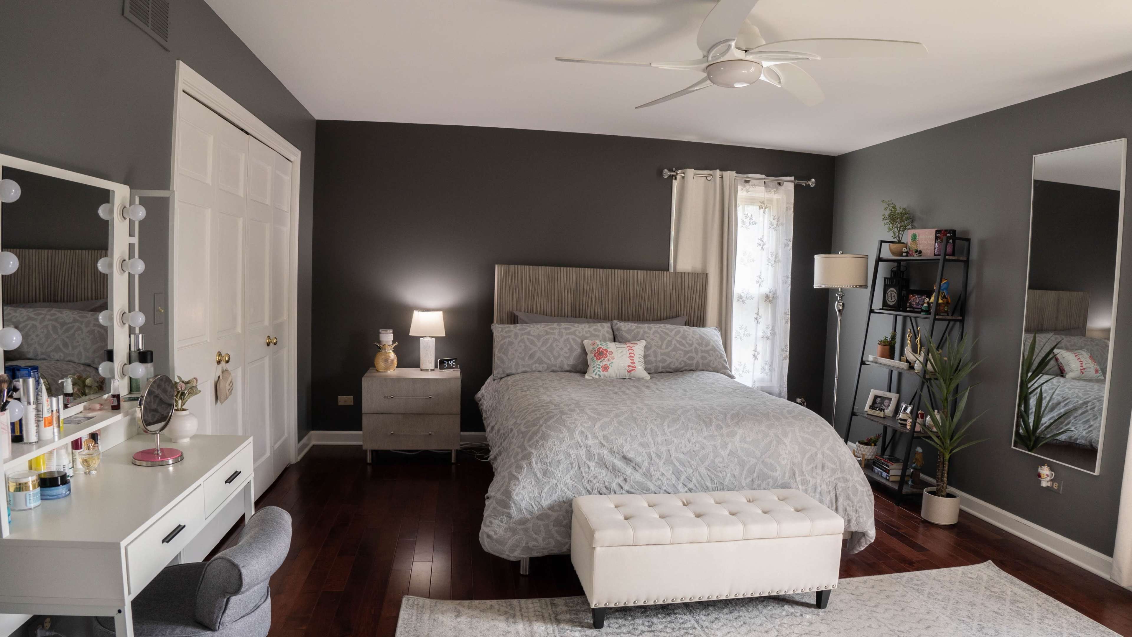 A modern bedroom with a queen-sized bed, a nightstand with a lamp, a standing mirror, and a small dressing table against a gray wall.
