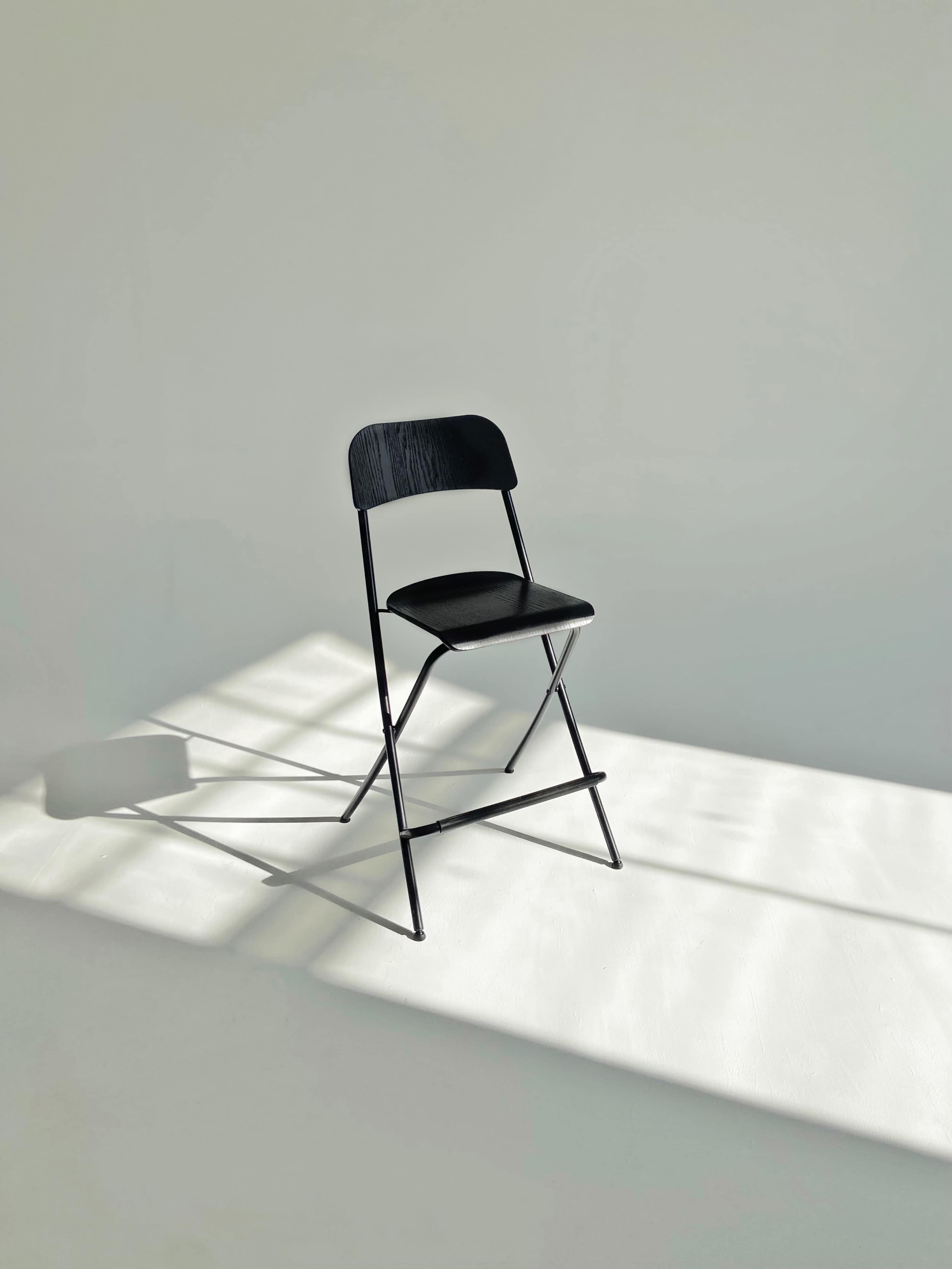A single black folding chair is positioned in a well-lit space, casting a shadow on the floor.