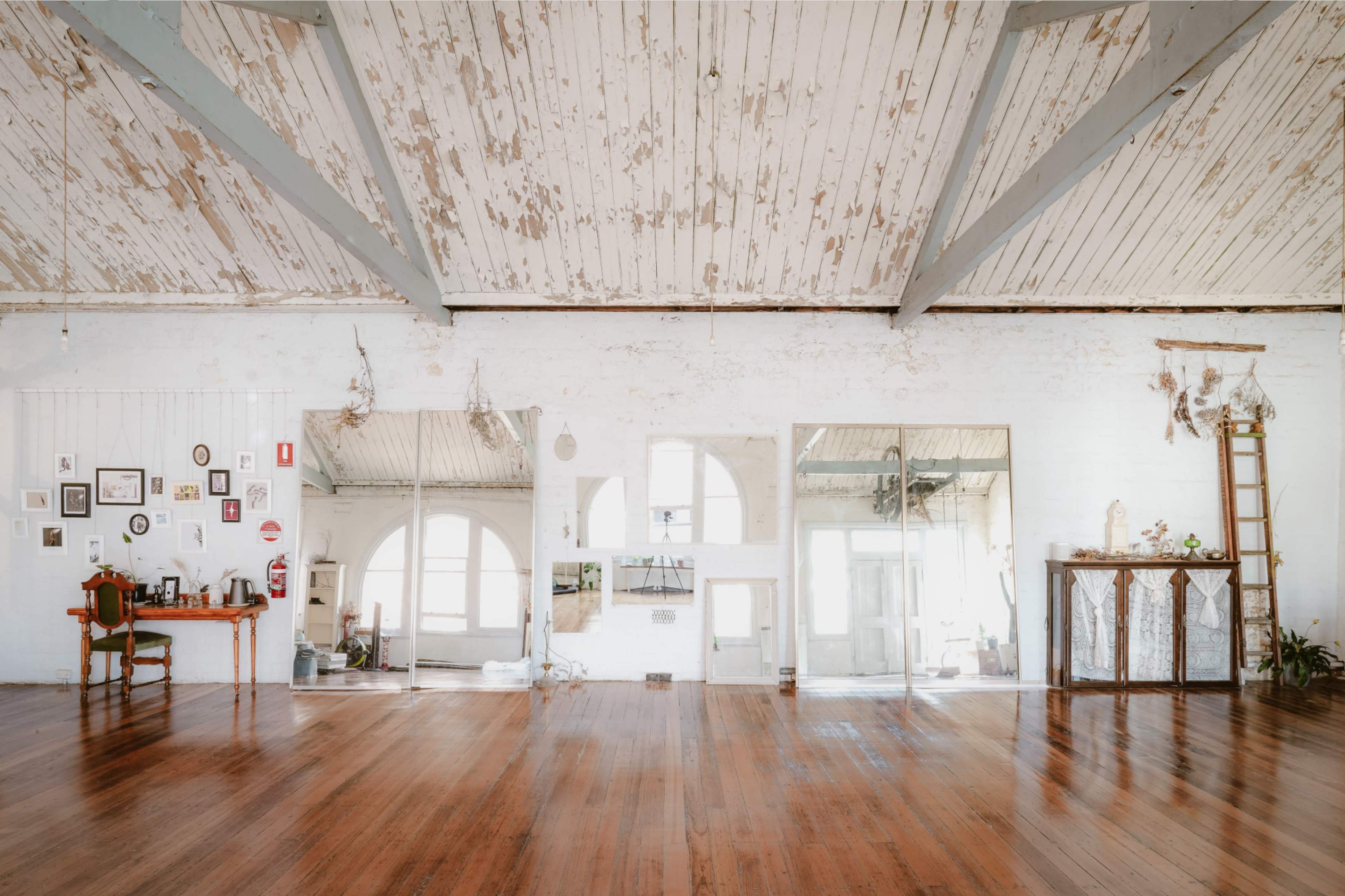 The image shows a spacious, brightly lit room with wooden floors, high ceilings, and large mirrors reflecting various decorative items and furniture.