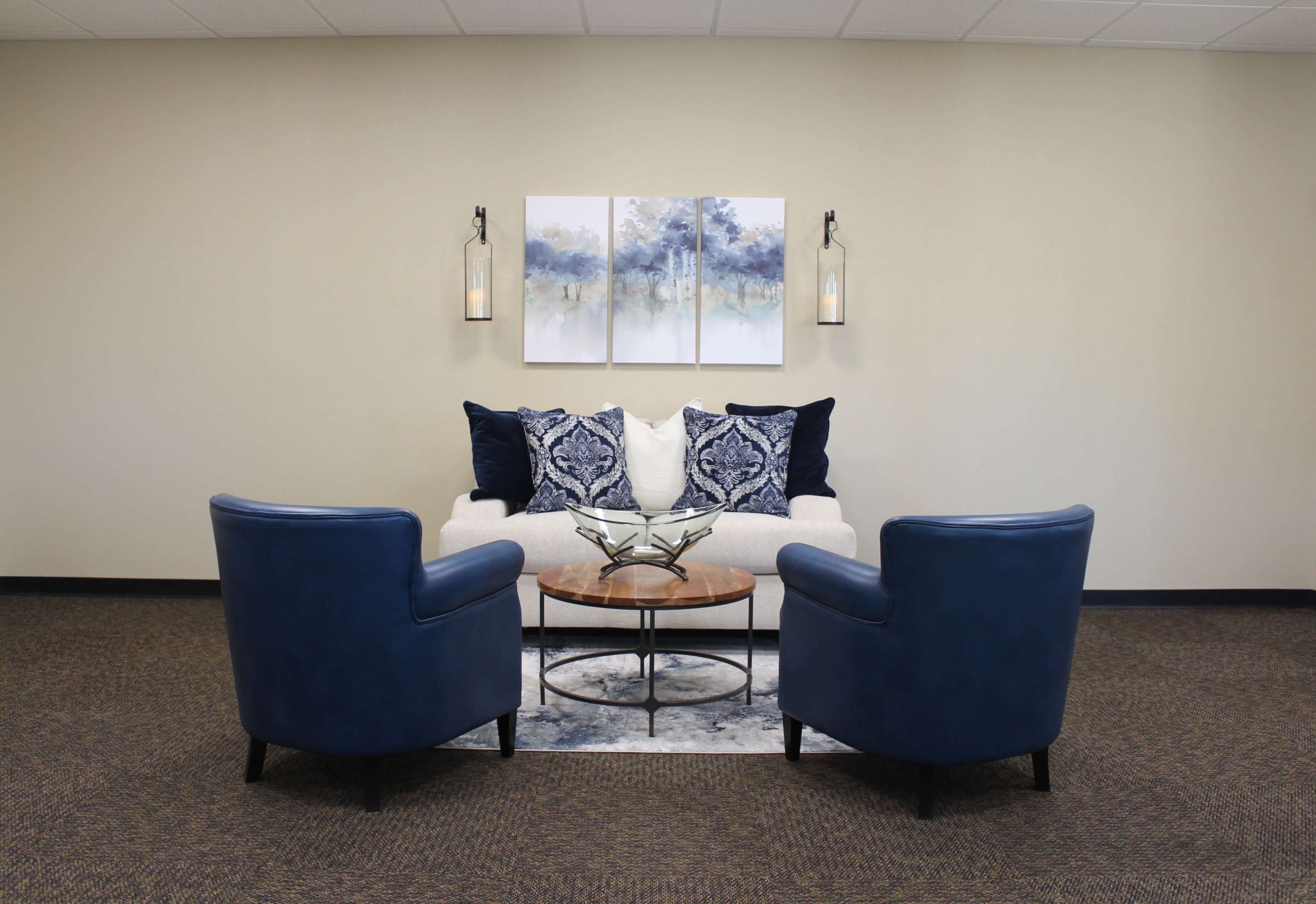 A cozy seating area featuring two blue armchairs facing a light-colored sofa, a round wooden coffee table, and wall art above the sofa.