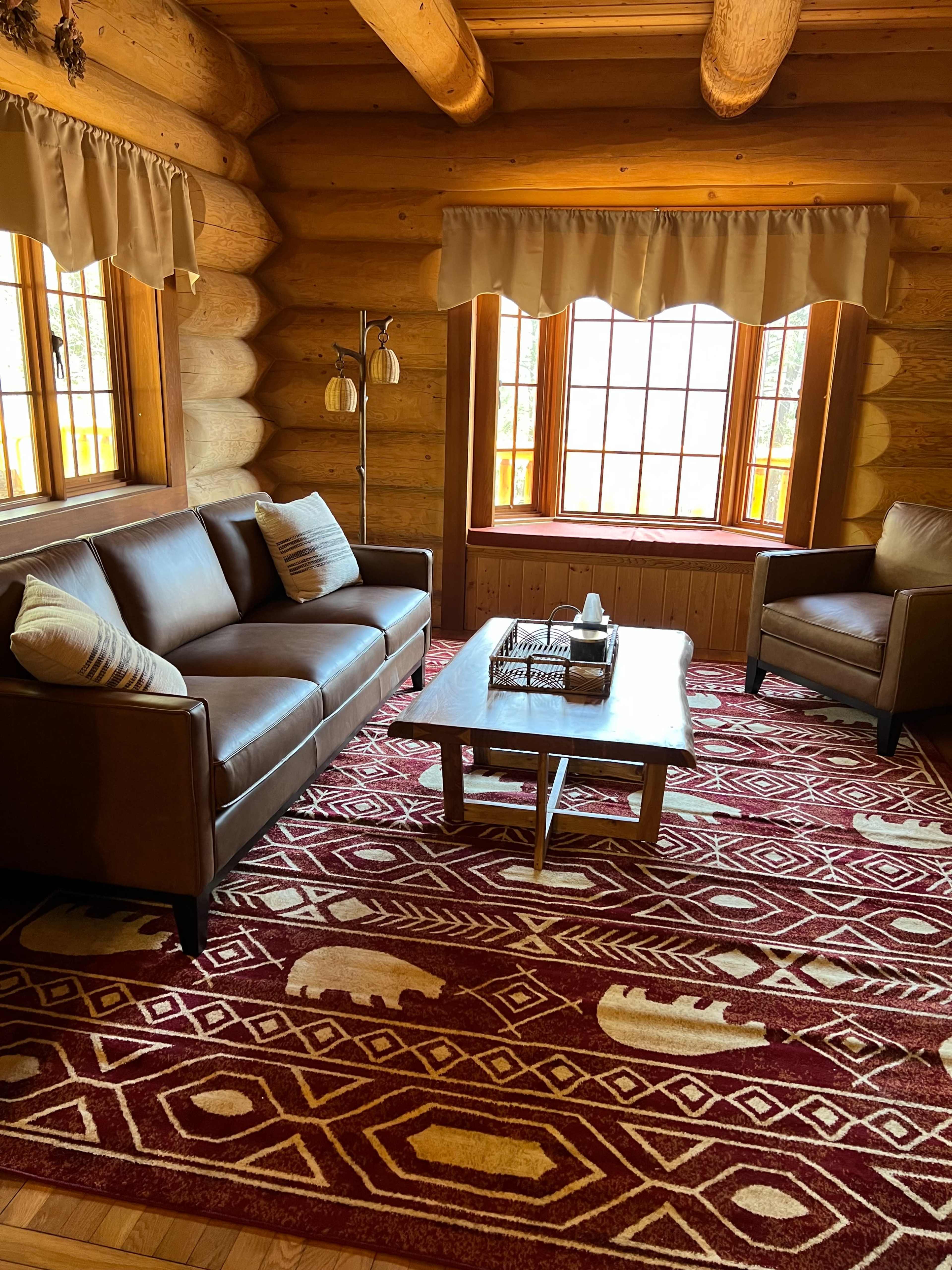 A rustic living room with log walls, a brown leather sofa and armchair, a wooden coffee table, and a patterned rug featuring bear motifs.