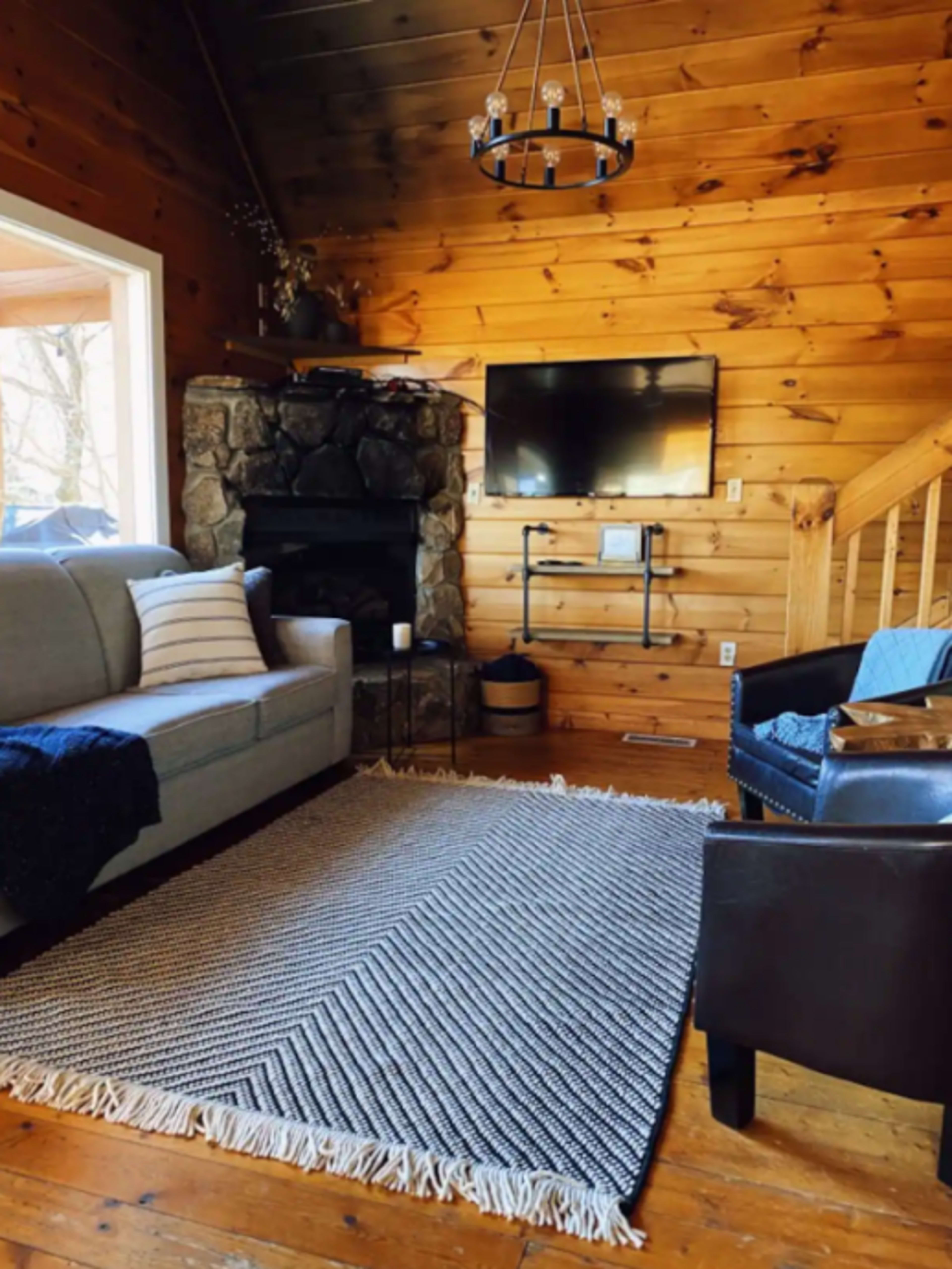 The image shows a cozy living room with a stone fireplace, a gray sofa, a black chair, a large area rug, and wooden walls.