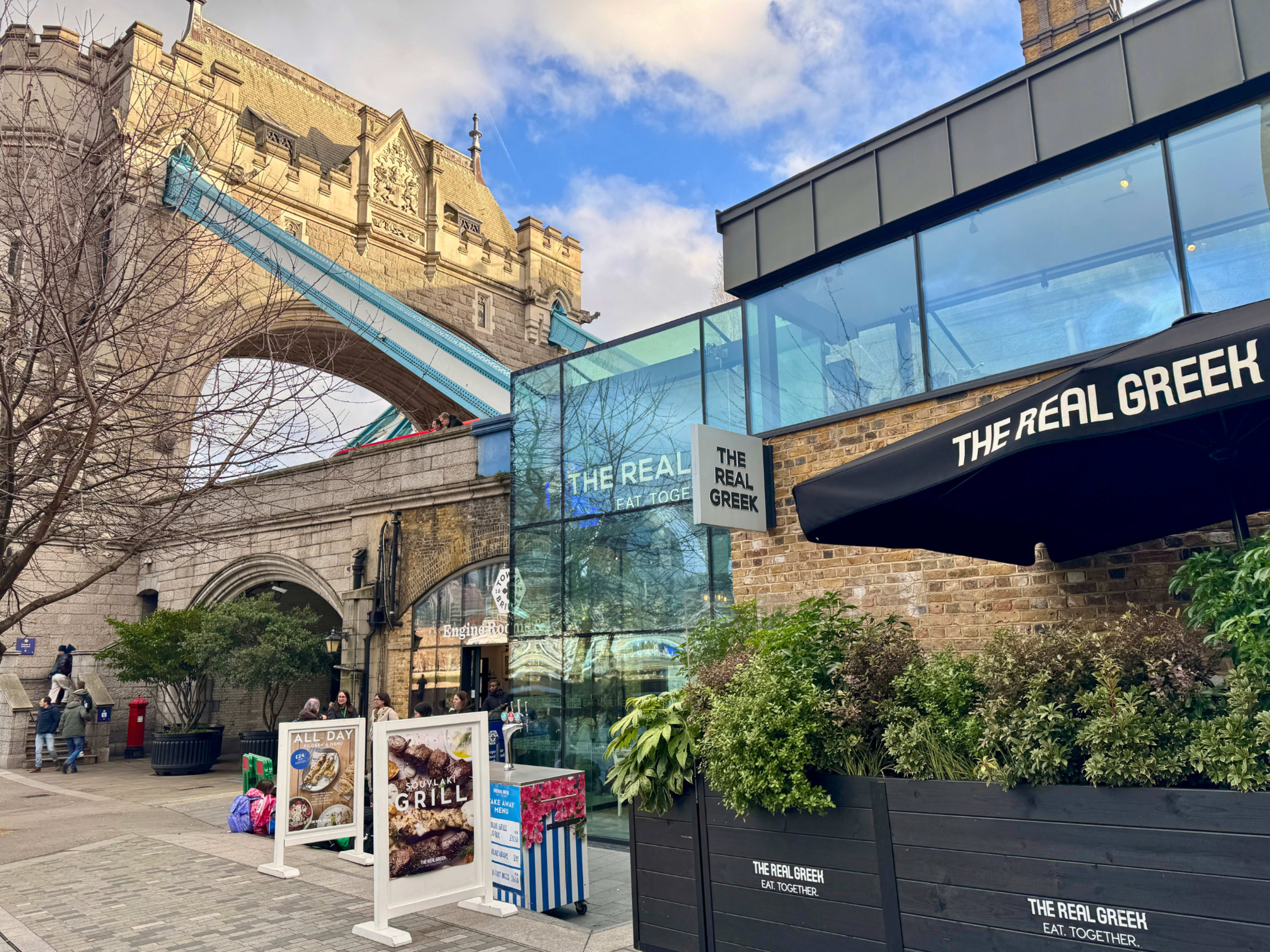 The image shows a restaurant called The Real Greek with outdoor seating, located near the Tower Bridge in London.