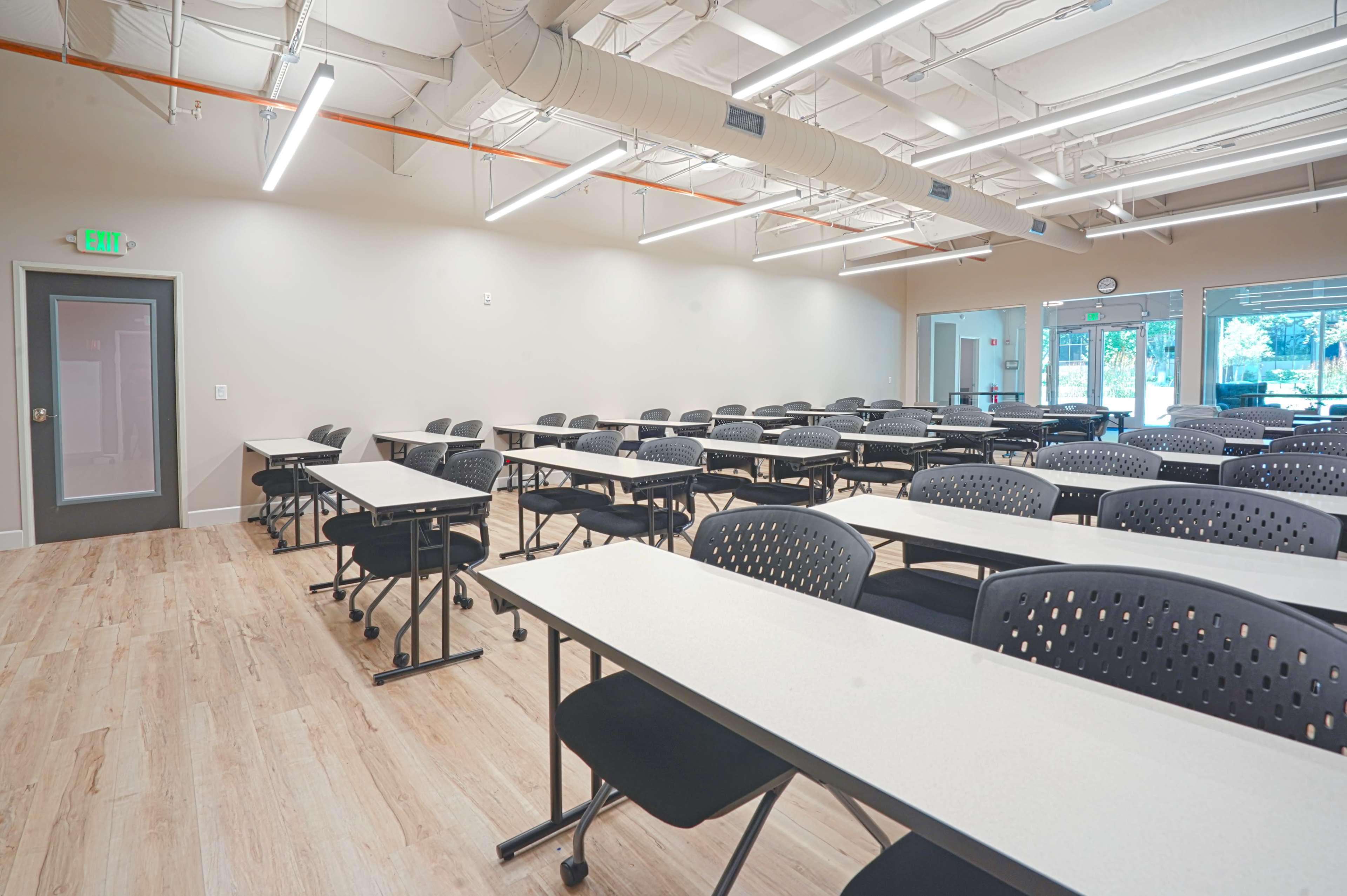 A spacious classroom features rows of tables and chairs arranged for student use, with large windows providing natural light.