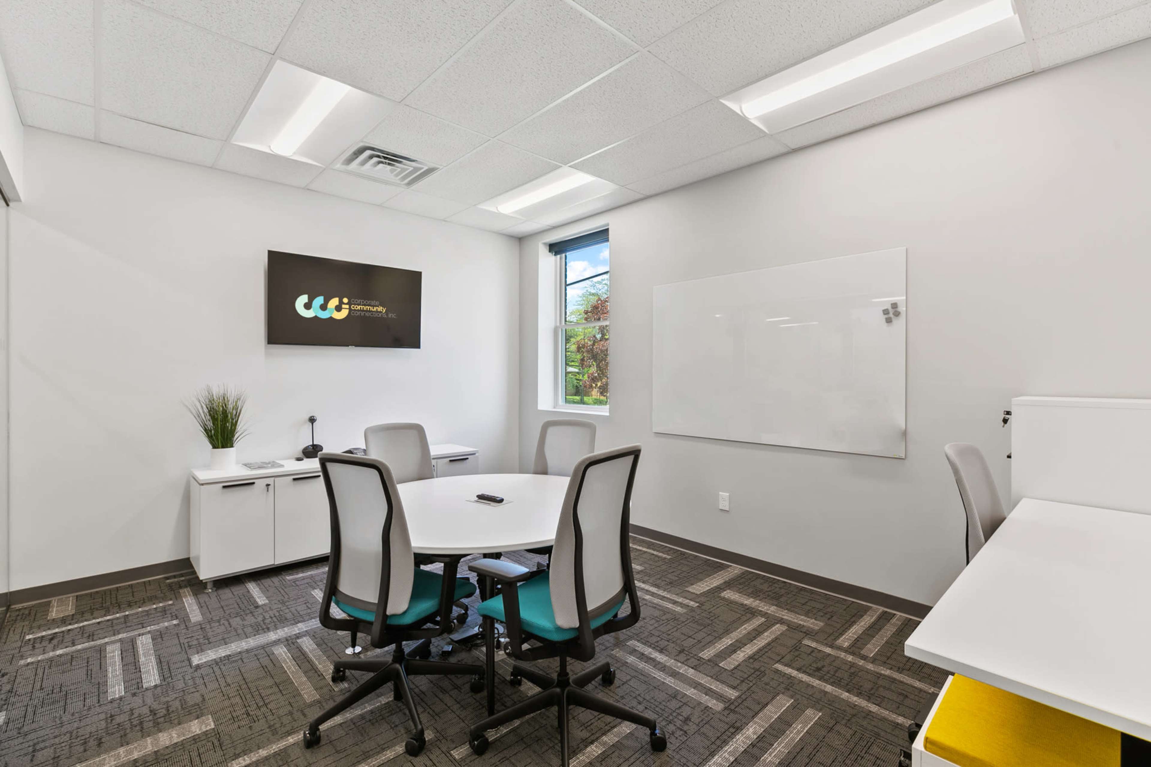 A modern conference room featuring a table surrounded by chairs, a wall-mounted screen, a whiteboard, and a window with a view outside.
