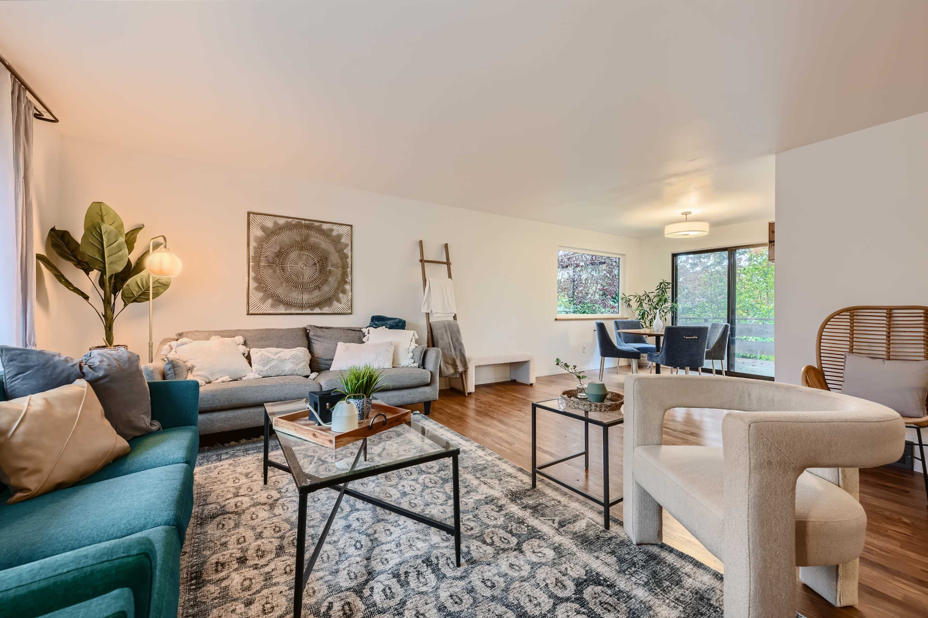 The image shows a modern living room with a green couch, a gray sofa, a glass coffee table, and large windows allowing natural light.