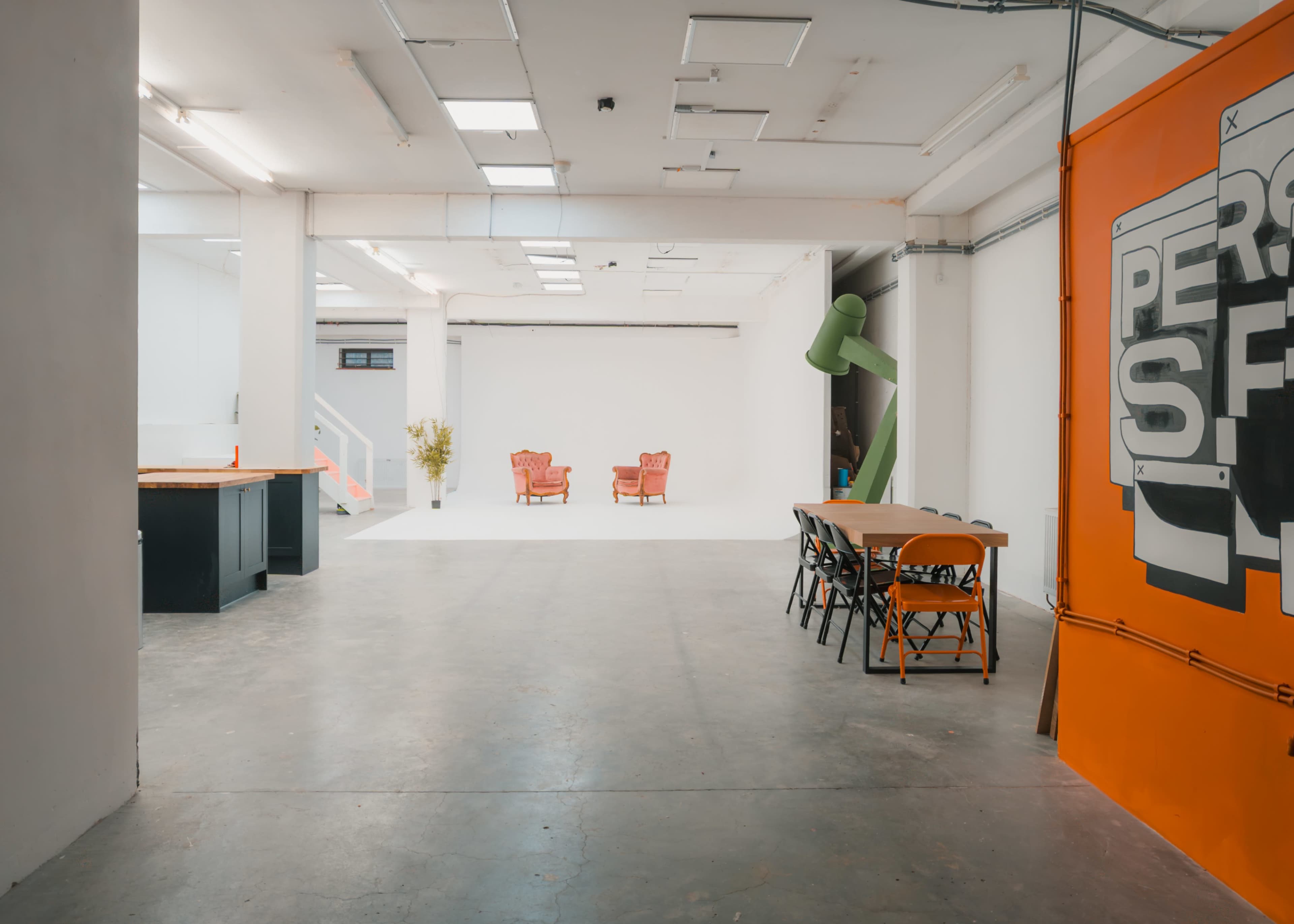 A large, minimalist studio space with two red chairs, a table set for dining, and a vibrant orange wall featuring text.