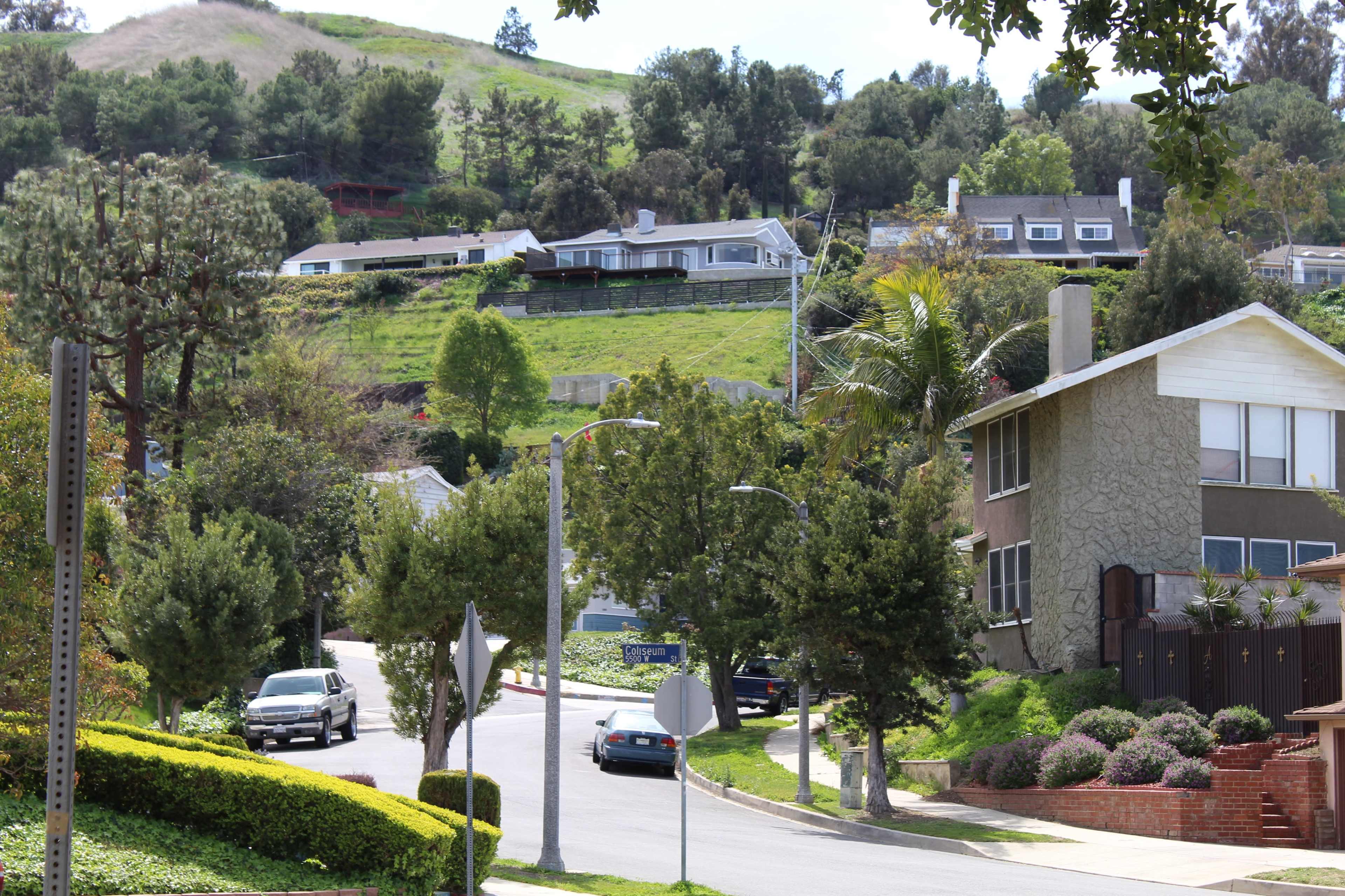 The image shows a residential neighborhood with houses situated on a hillside, surrounded by greenery and trees.