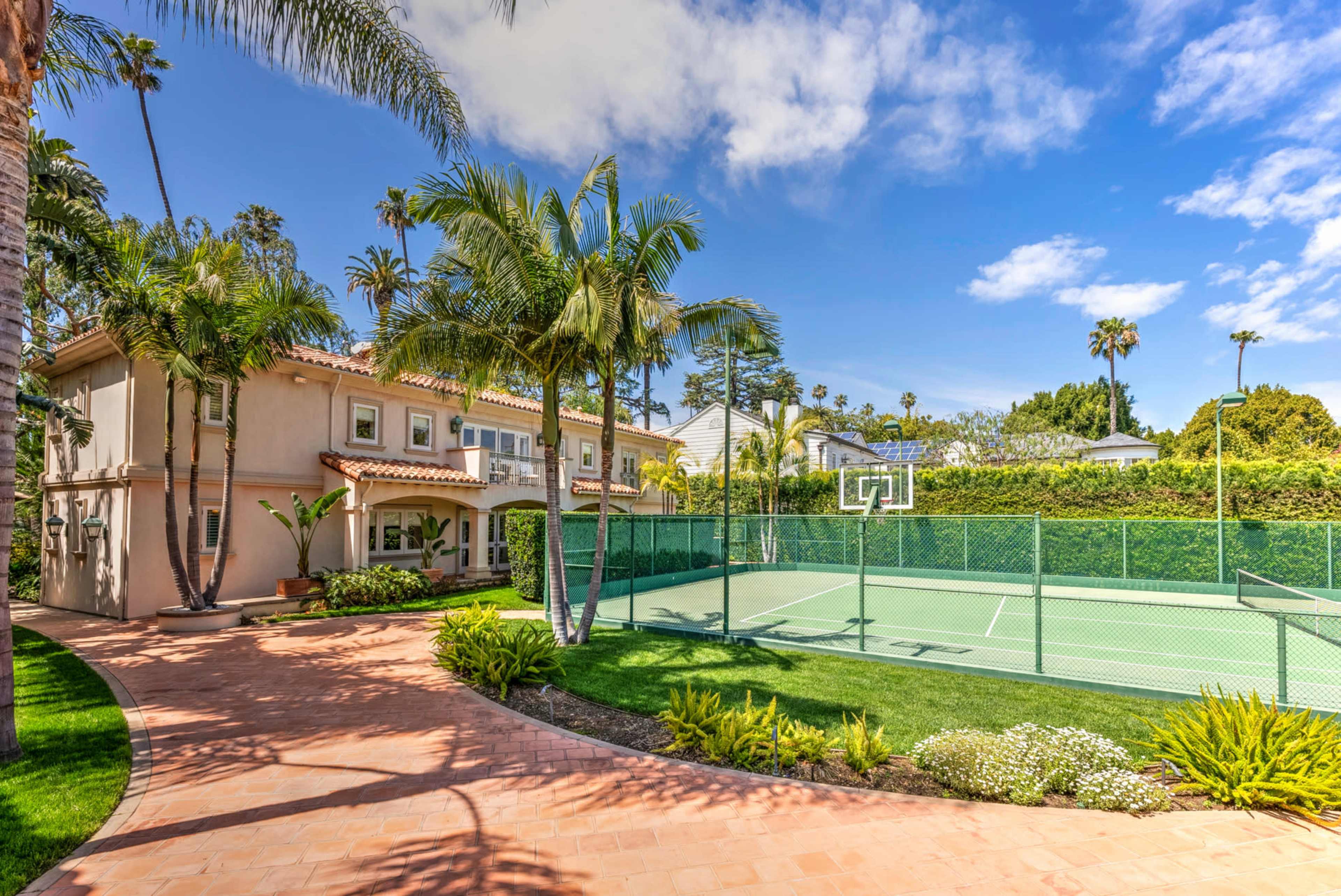 The image shows a tennis court surrounded by a landscaped yard and a two-story house in a sunny residential area with palm trees.