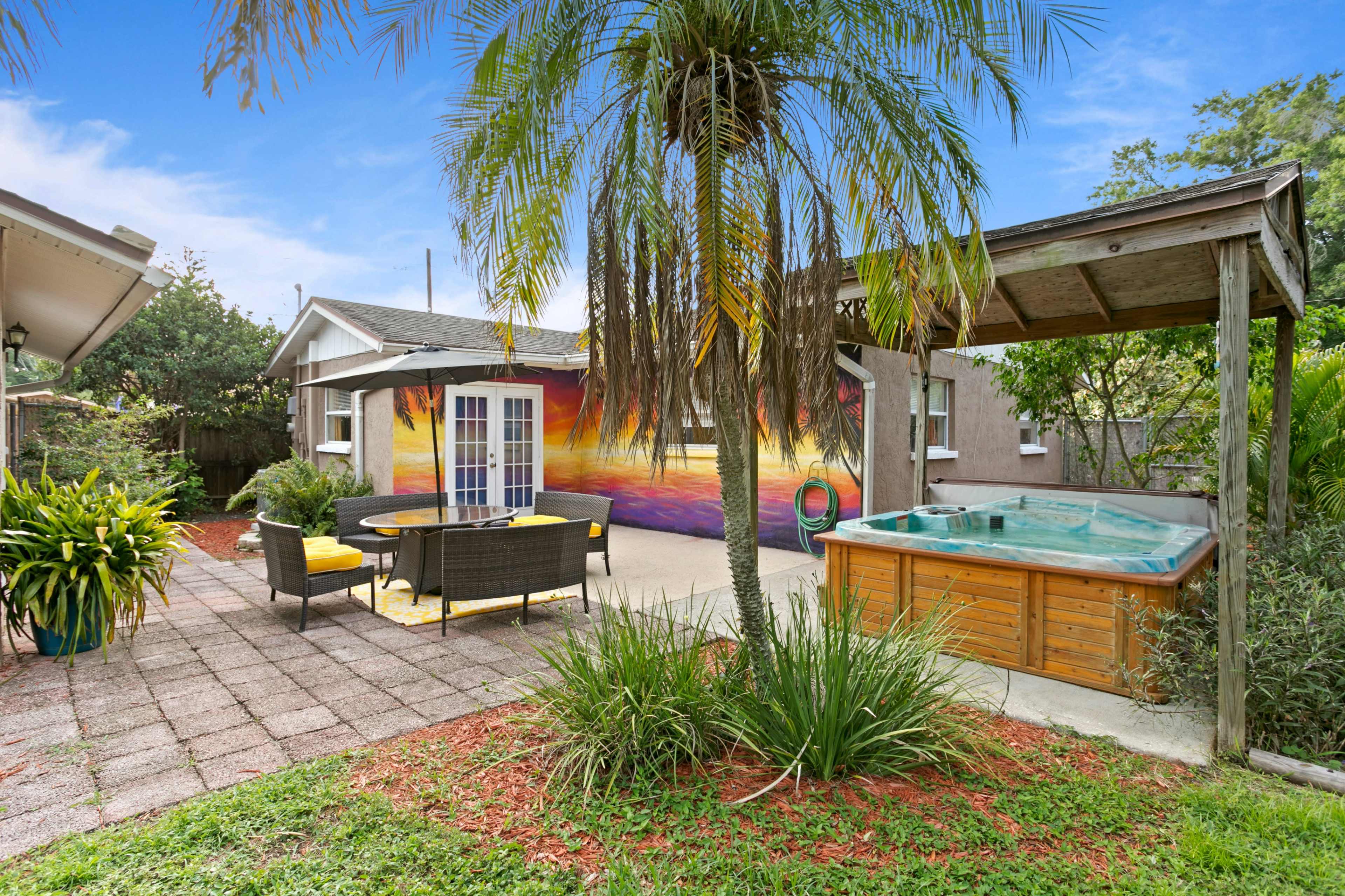 A backyard patio with a dining table and chairs, a hot tub, a colorful mural on the wall, and surrounding greenery.