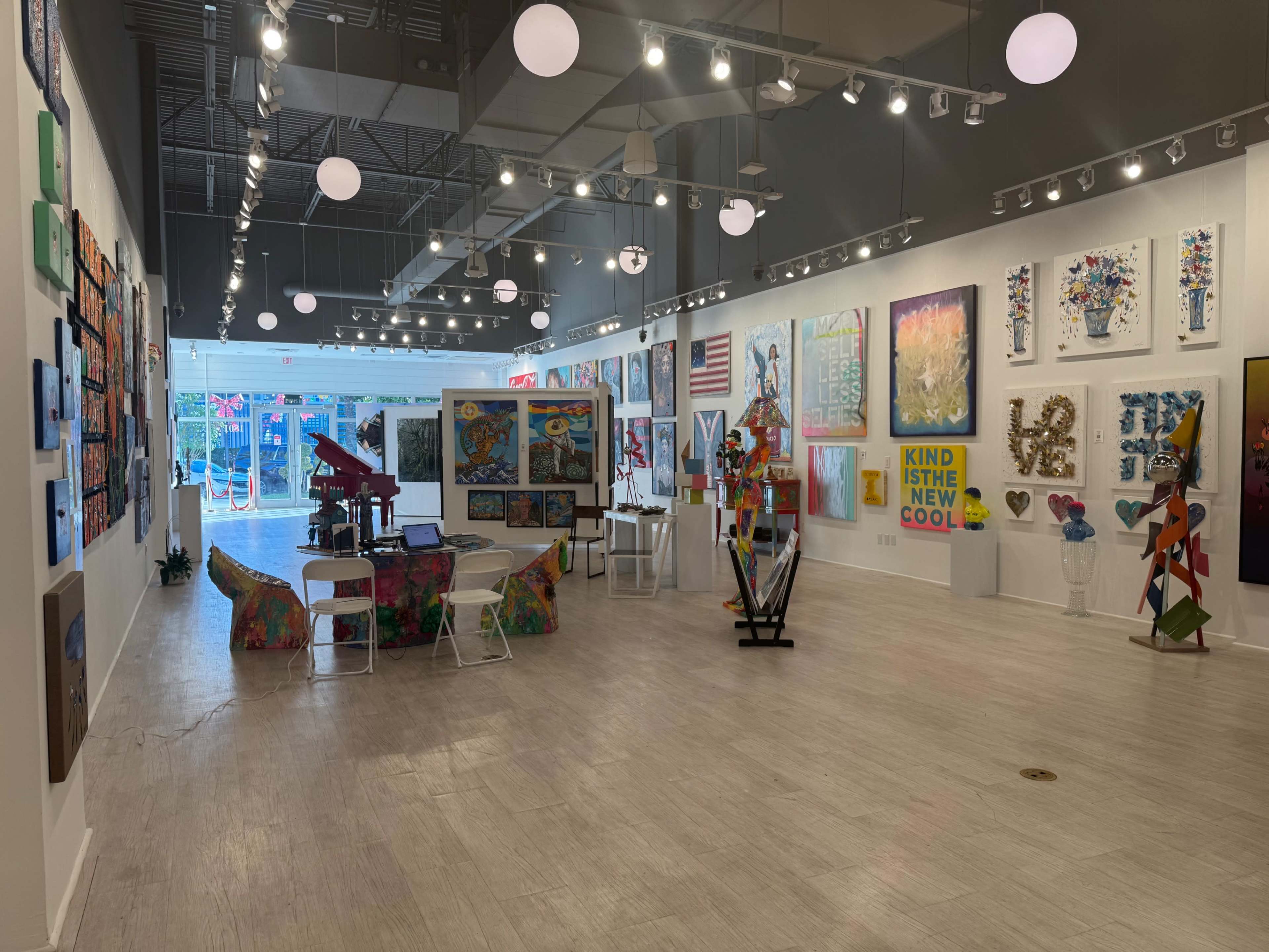 The image shows an art gallery with colorful artworks displayed on white walls, featuring a range of sculptures and seating arrangements throughout the space.