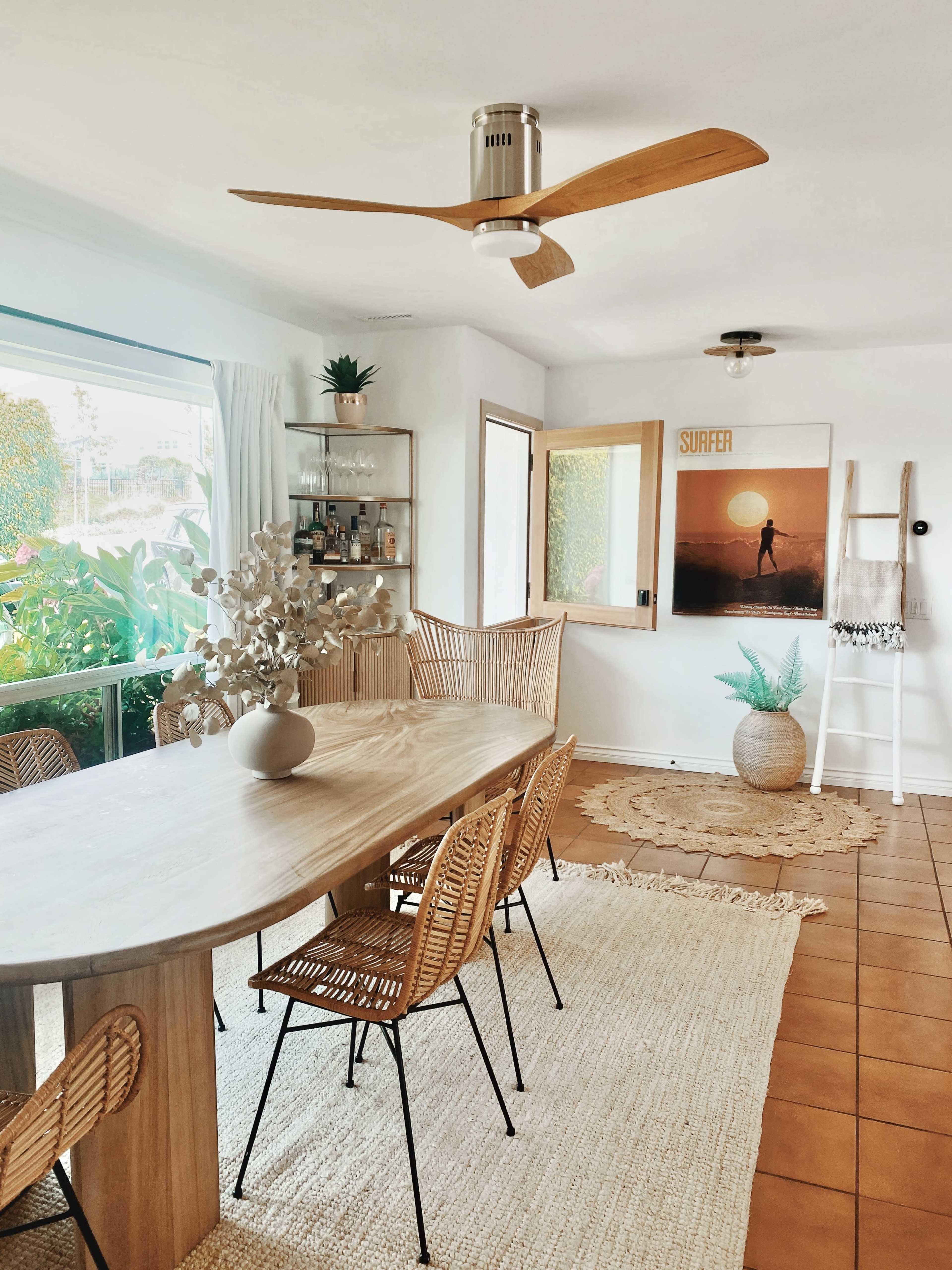 A well-lit dining area featuring a wooden table surrounded by rattan chairs, a ceiling fan, and decorative elements like plants and wall art.