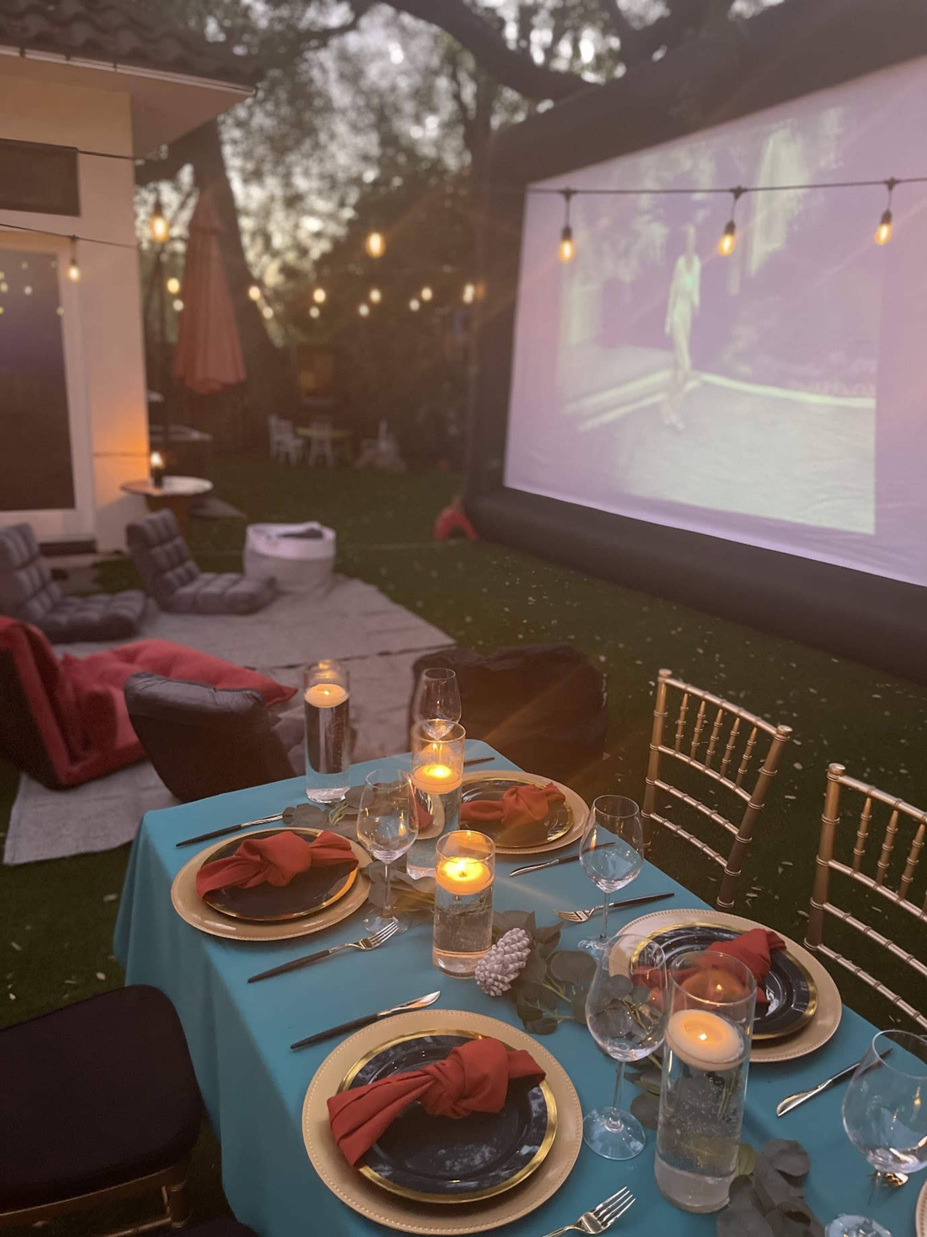 A beautifully set outdoor dining table with turquoise tablecloth, elegant tableware, and candles, alongside a large inflatable screen showing a film in a backyard illuminated by string lights.