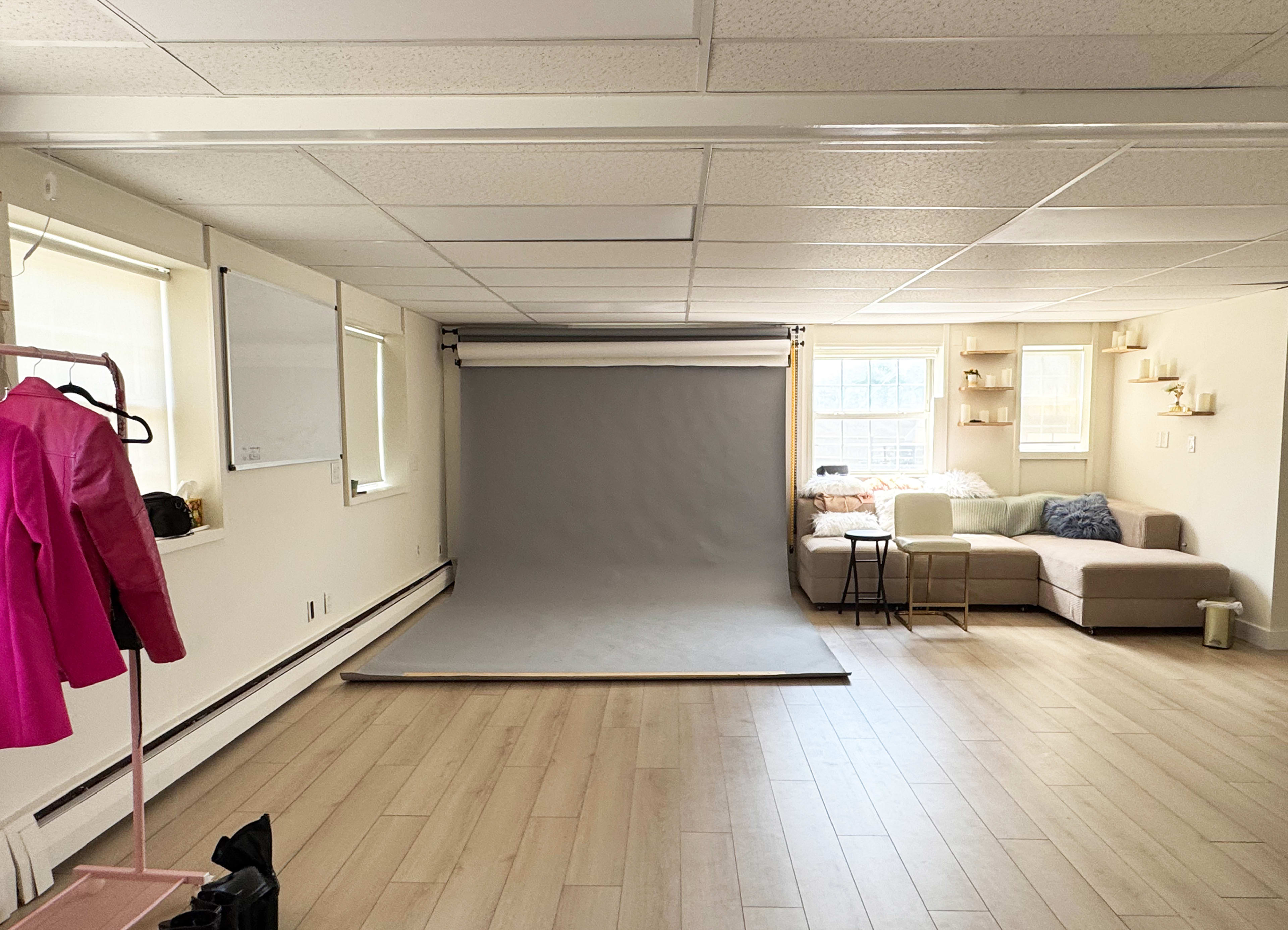 The image shows an interior room with light wooden flooring, featuring a photography backdrop, a couch, and a clothing rack with a pink jacket.