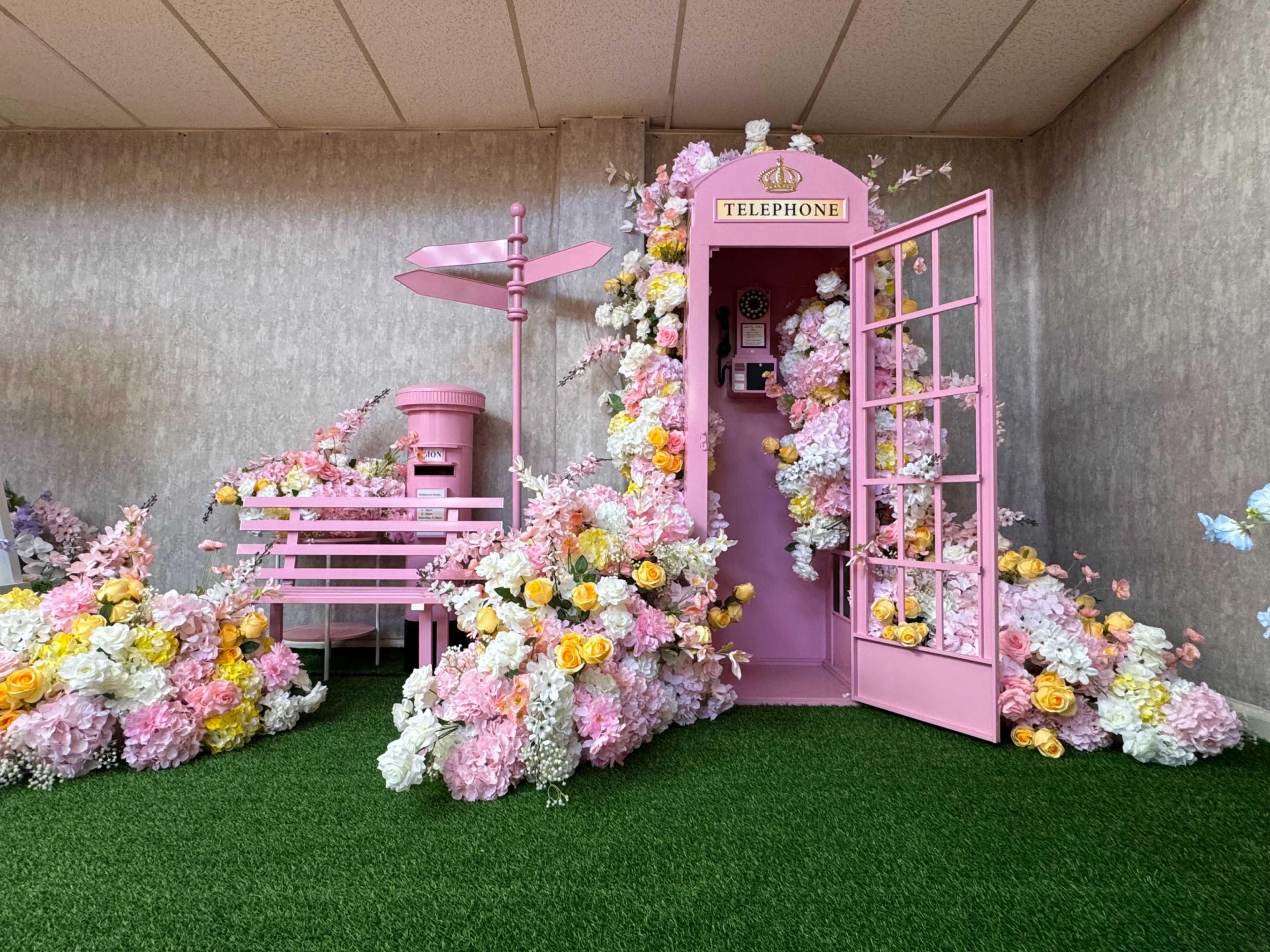 The scene features a vibrant pink telephone booth surrounded by an arrangement of artificial flowers, with a matching pink mailbox and a decorative fence in a grassy area.