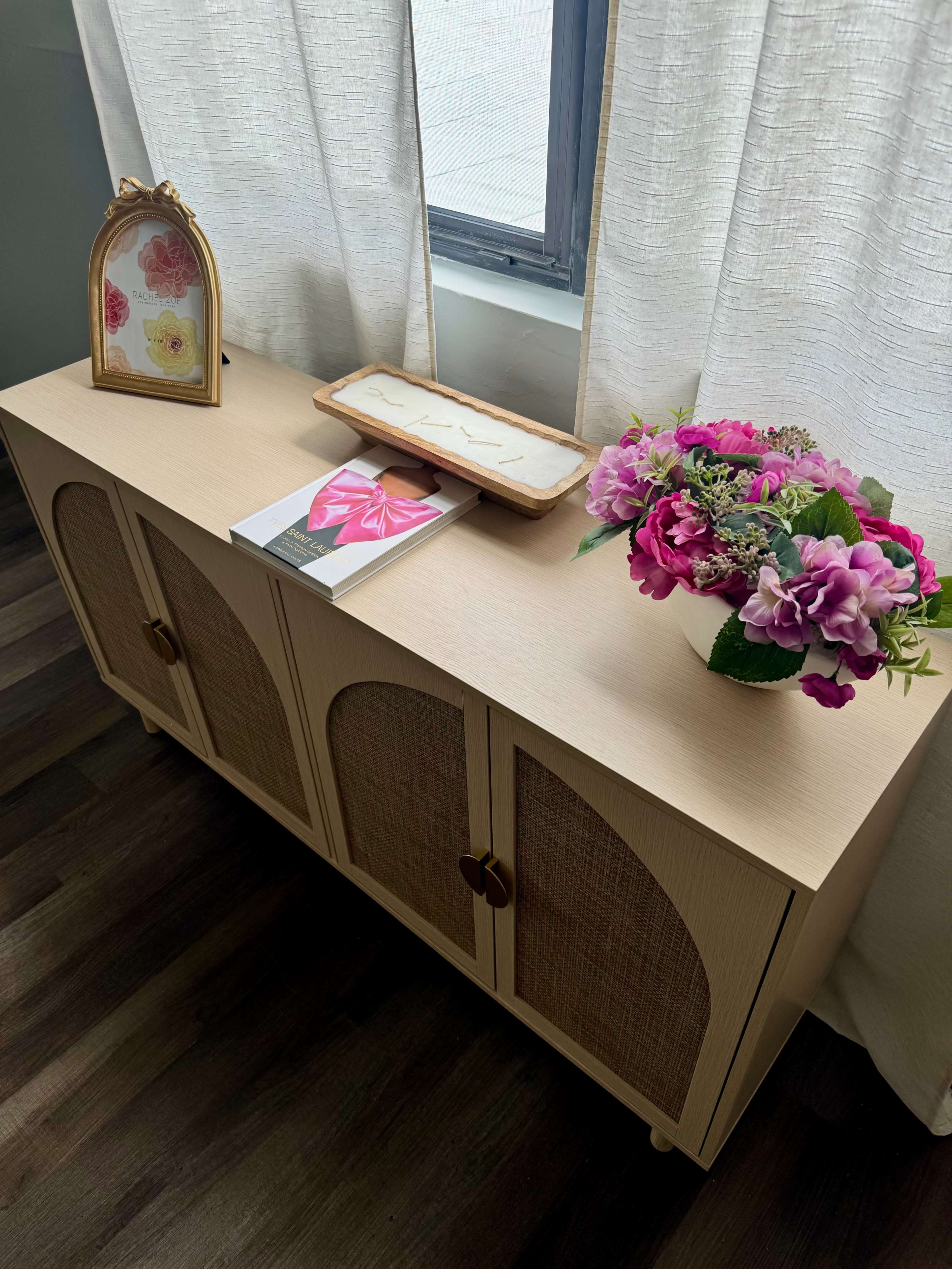 A light-colored cabinet with decorative doors sits against a wall, featuring a floral arrangement, a photo frame, and a small wooden tray on top.