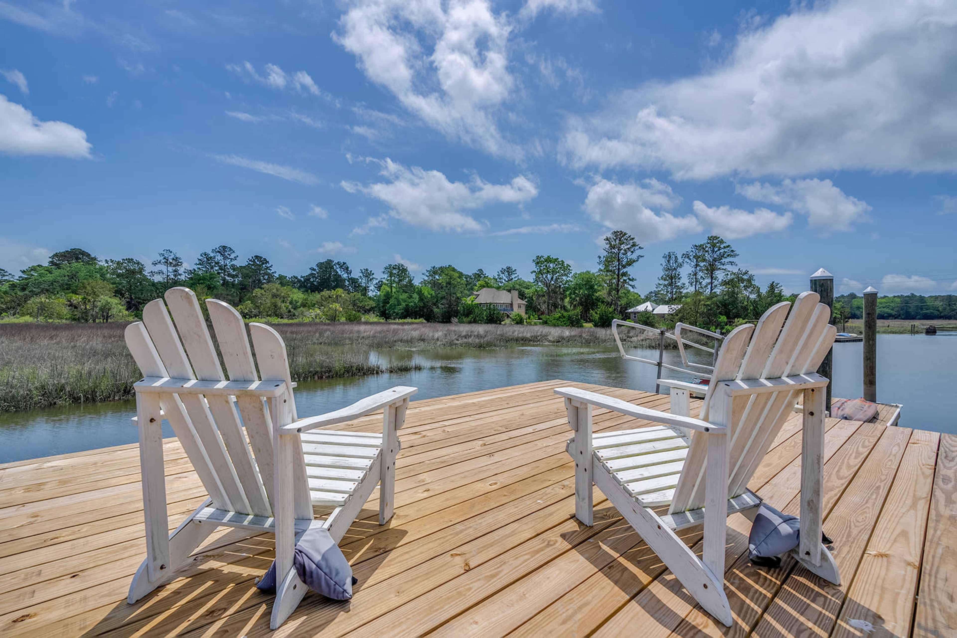 Two white Adirondack chairs are positioned on a wooden dock overlooking a calm body of water surrounded by greenery.