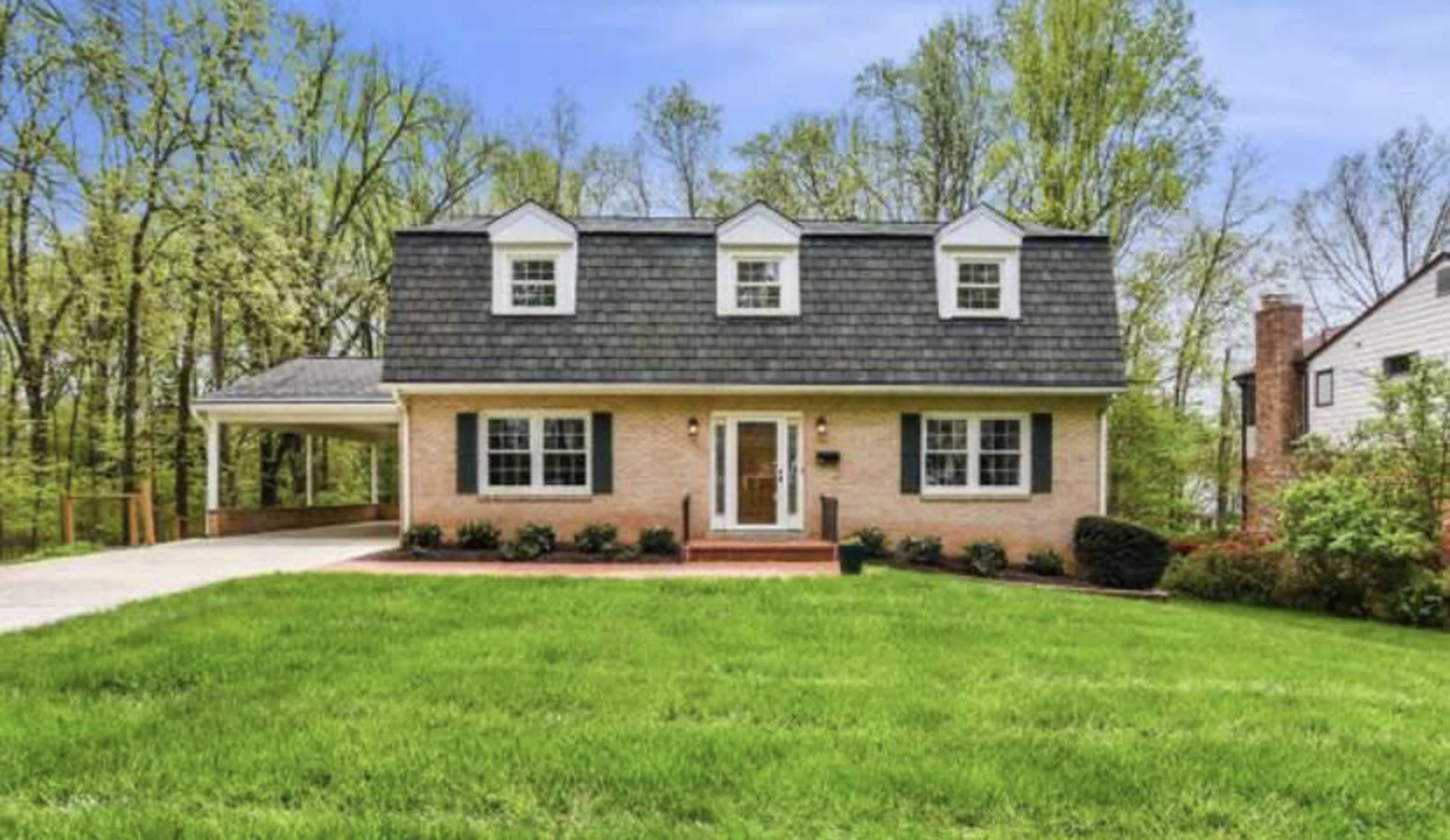 A two-story house with a gray shingles roof and a brick exterior is set on a grassy lawn surrounded by trees.
