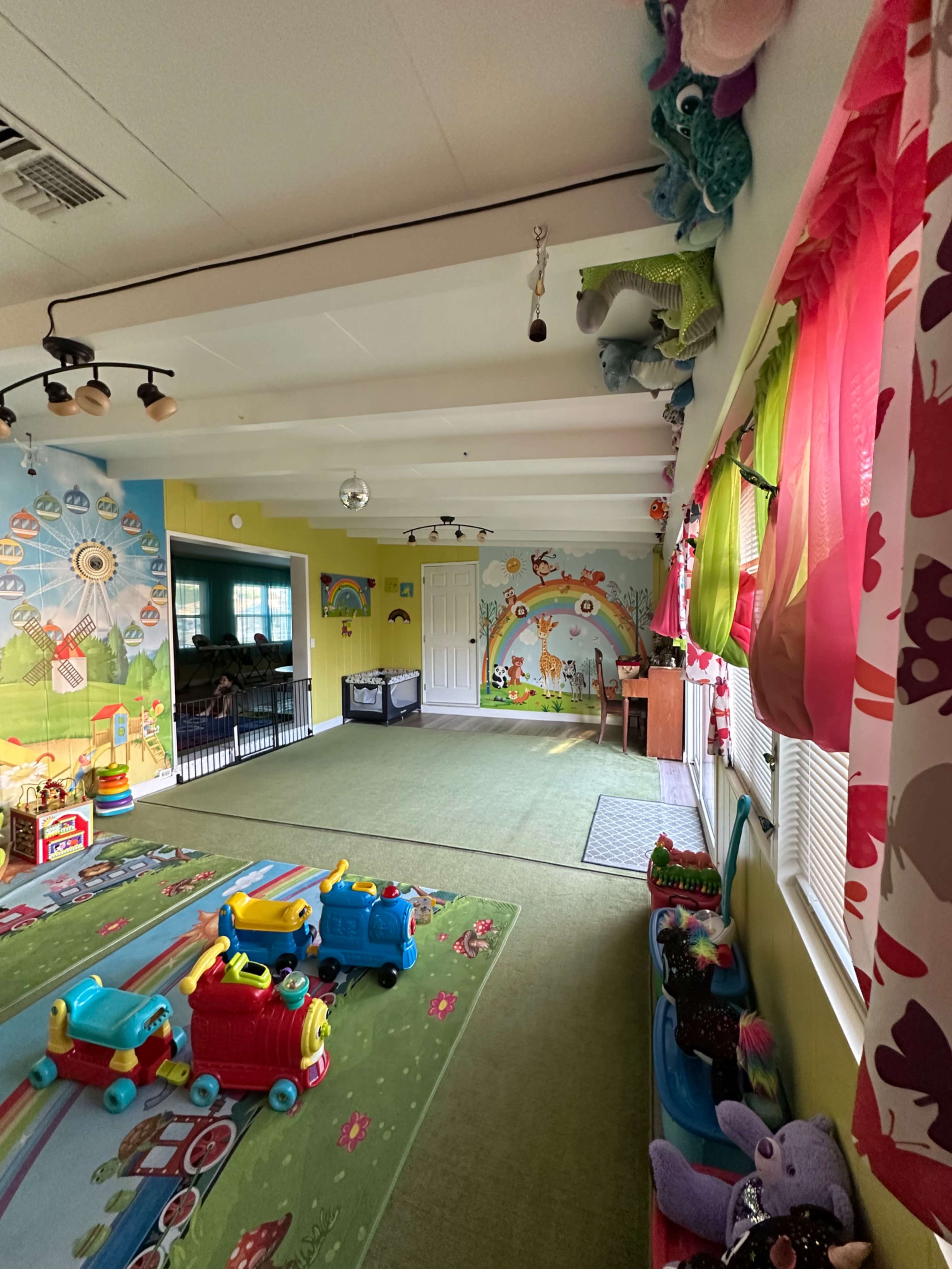 The image shows a colorful playroom with toy trains on a green carpet, decorated walls featuring murals, and large windows covered with bright curtains.