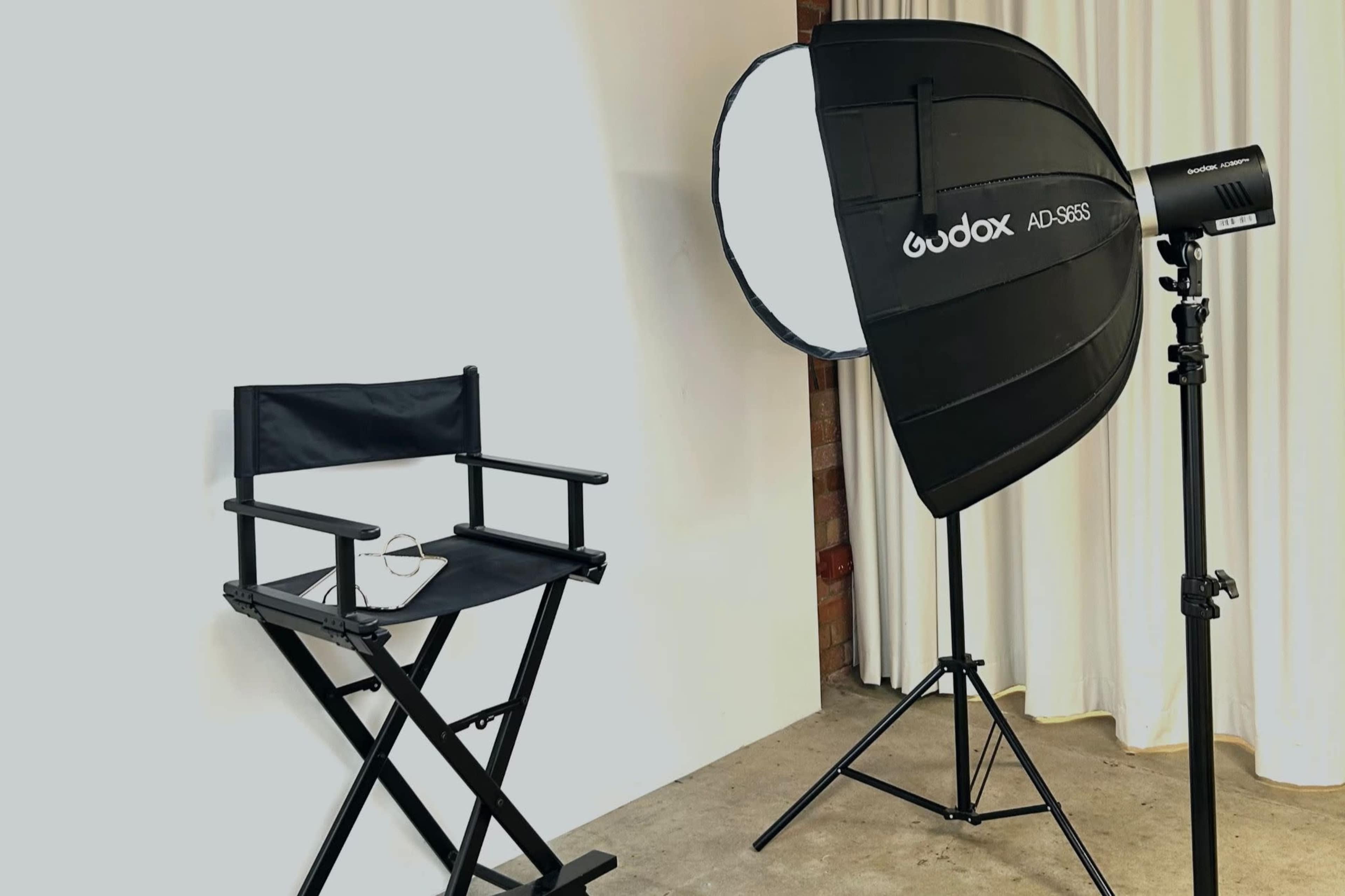 A photography setup with a director's chair beside a large softbox light on a stand, positioned against a backdrop of curtains.
