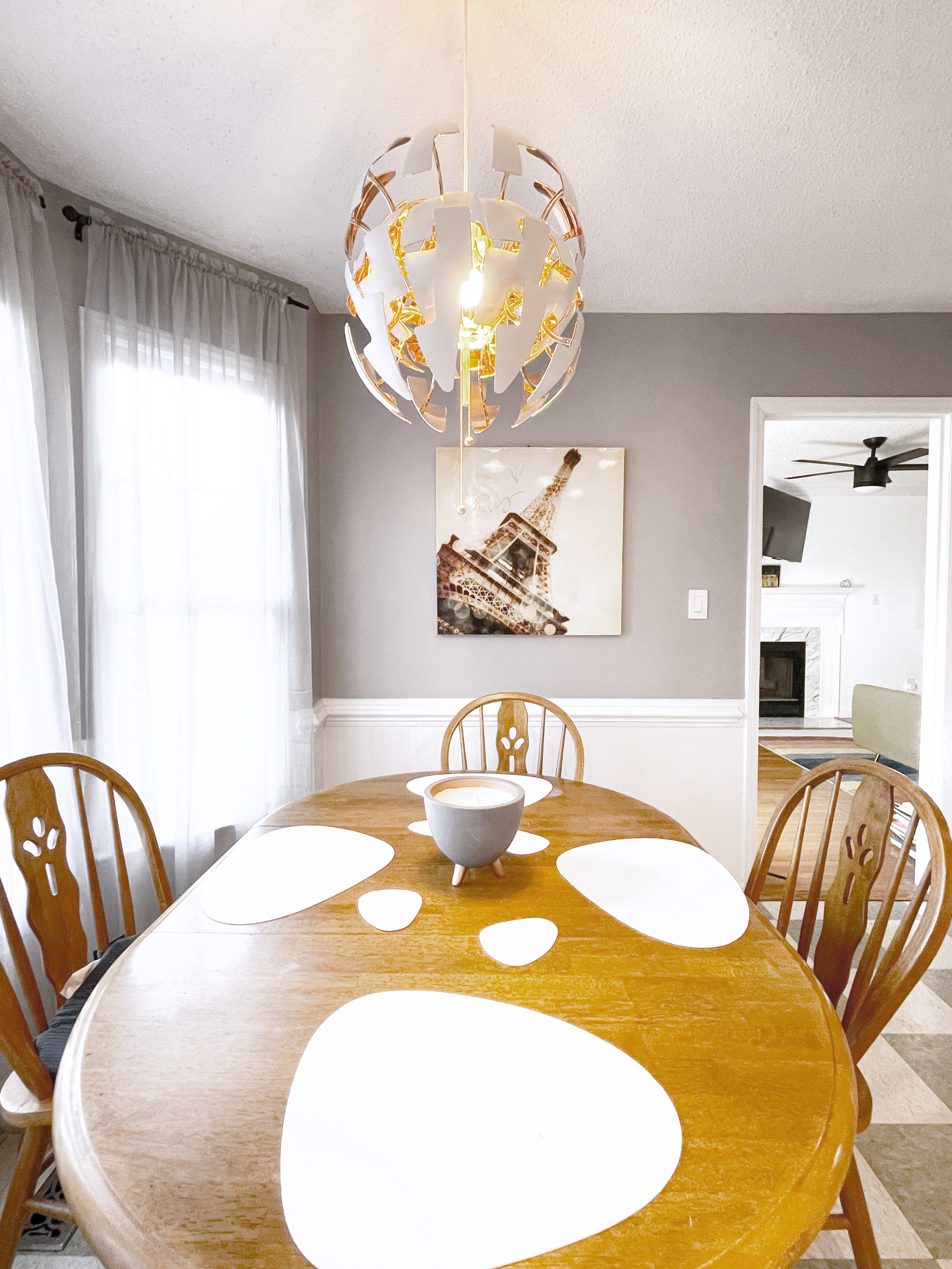 A wooden dining table set with white placemats and a bowl sits beneath a decorative light fixture in a room featuring a wall art piece of the Eiffel Tower.