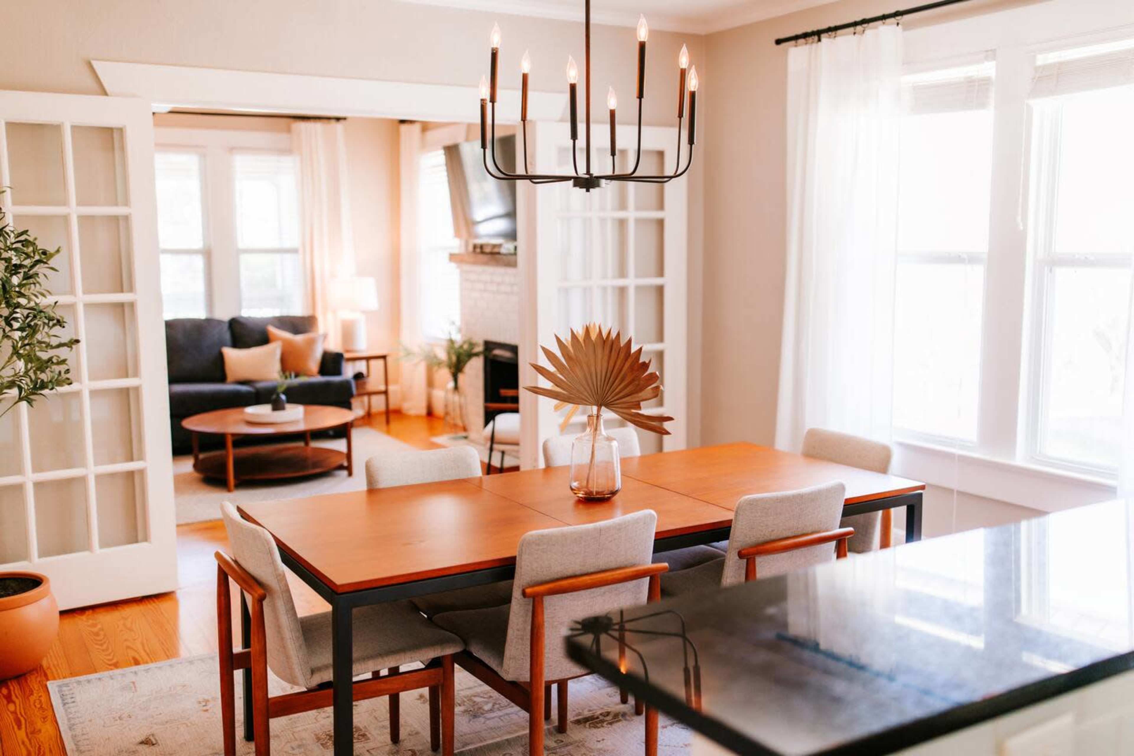 The image shows a dining area with a wooden table surrounded by six chairs, a chandelier above, and a view into a living room with a sofa and coffee table.